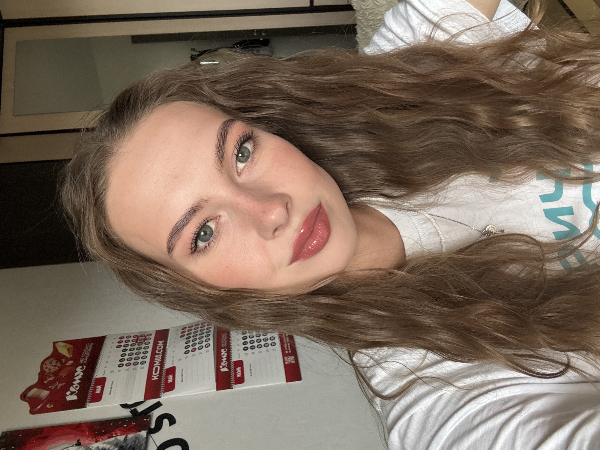 A young female adult, appearing to be in her late teens or early twenties, is depicted in a close-up selfie, looking directly at the camera. She has long, wavy, light brown hair, light green eyes, and is wearing a white t-shirt with faint blue lettering visible on the chest. A silver necklace with a heart-shaped pendant is worn around her neck. Her face features light makeup, including defined eyebrows, mascara, blush, and glossy coral-pink lipstick.
The indoor background on the left reveals a white wall adorned with a partially visible dark poster featuring white text and a red element. Below this is a red and white tear-off calendar, prominently displaying the Russian brand "Комус" (Komus) and "KOMUS.COM". Visible calendar sheets include "МАЙ" (May) and "ИЮНЬ" (June), showing numbered days from 19 to 26 with circular markings. On the right side of the frame, a mirrored wardrobe or door reflects an indistinct room interior, and a section of fluffy, light-colored fabric is visible at the bottom right. The presence of the Komus calendar, a Russian office supply company, and Cyrillic text strongly indicates the location is in Russia, specifically consistent with the city of Perm where Komus operates.