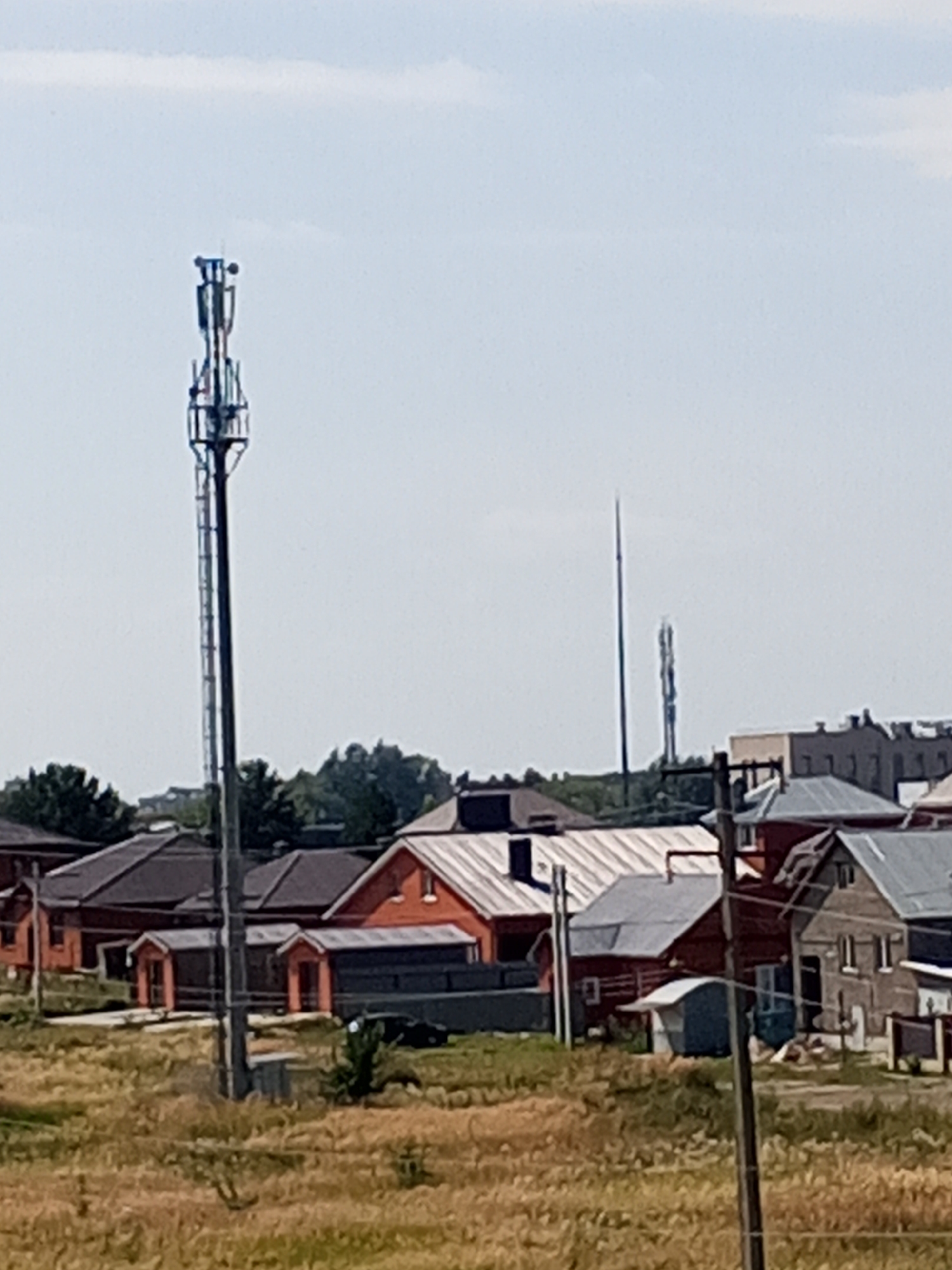 A cellular tower stands prominently in the foreground against a light blue sky with scattered clouds. The tower, made of metal lattice, has multiple antennas and equipment at its apex. In the midground, a collection of residential buildings with pitched roofs is visible. Some of these buildings are constructed with brick, while others appear to have darker roofing materials. The area in front of the buildings is covered in dry, yellow grass. Further in the background, another, more slender tower is visible, along with more buildings and trees. The scene suggests a suburban or rural setting on a clear day. The image does not contain any people or discernible actions.
