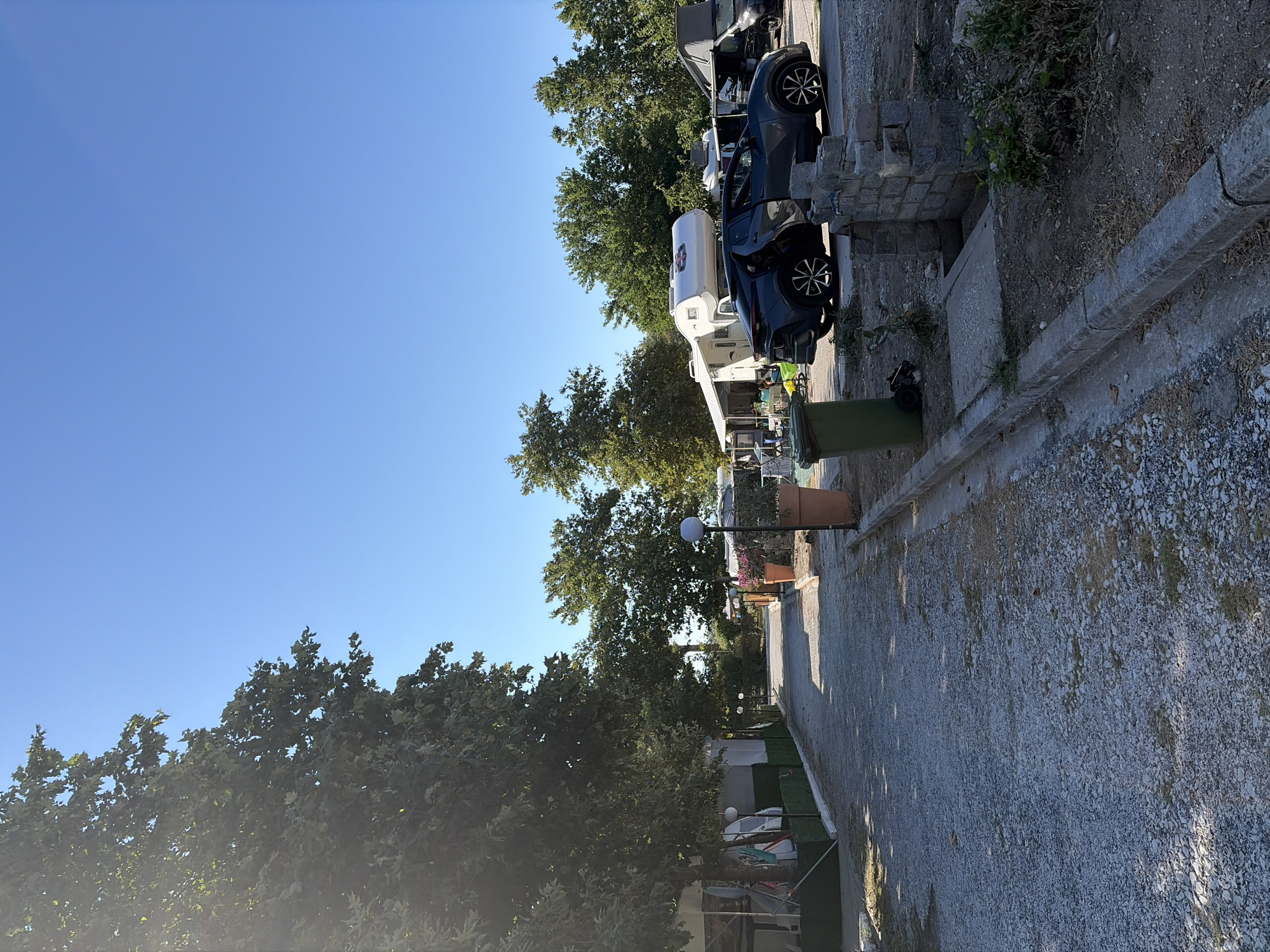 This image depicts a campsite scene under a clear blue sky. In the foreground, a gravel path runs through the campsite, bordered by a concrete curb and dirt. On the left side of the path, there are white structures resembling temporary accommodations or stalls, with some laundry hanging. Large trees with dense green foliage cast shadows on the path.
Further down the path, on the right side, large terracotta pots are visible, some containing plants. A dark-colored SUV with its doors open is parked in the middle ground. Behind the SUV, a white camper van is parked next to a white awning. Further in the background, more trees and structures suggesting other camping units are present. A tall green trash bin is located near the SUV.
The overall environment suggests a sunny day in a relaxed outdoor setting, likely a campsite, possibly located in or near Kavala, Greece, given the context of the prompt. The image is shot from a slightly low angle, looking down the path. There are no clearly visible individuals engaged in any specific actions.