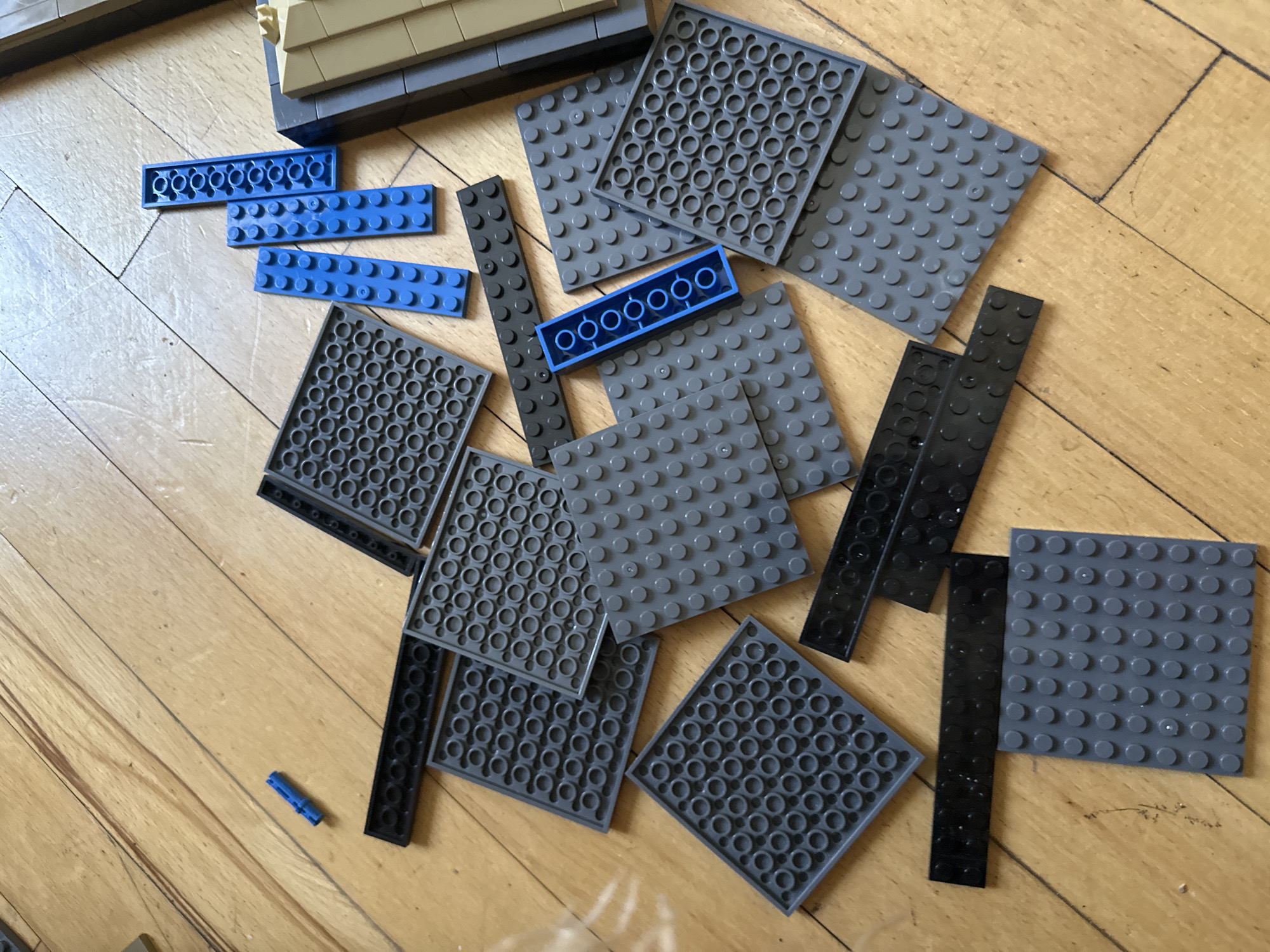 A collection of scattered LEGO bricks, primarily gray and blue, are spread across a light brown wooden floor. The LEGO pieces include flat base plates of various sizes and some smaller building bricks. The lighting suggests an indoor environment, possibly a room with natural light. The image is a close-up, focusing on the arrangement of the LEGOs, and does not depict any urban elements or provide any geographical indicators that would suggest a location in Yerevan, Armenia, or any other city.