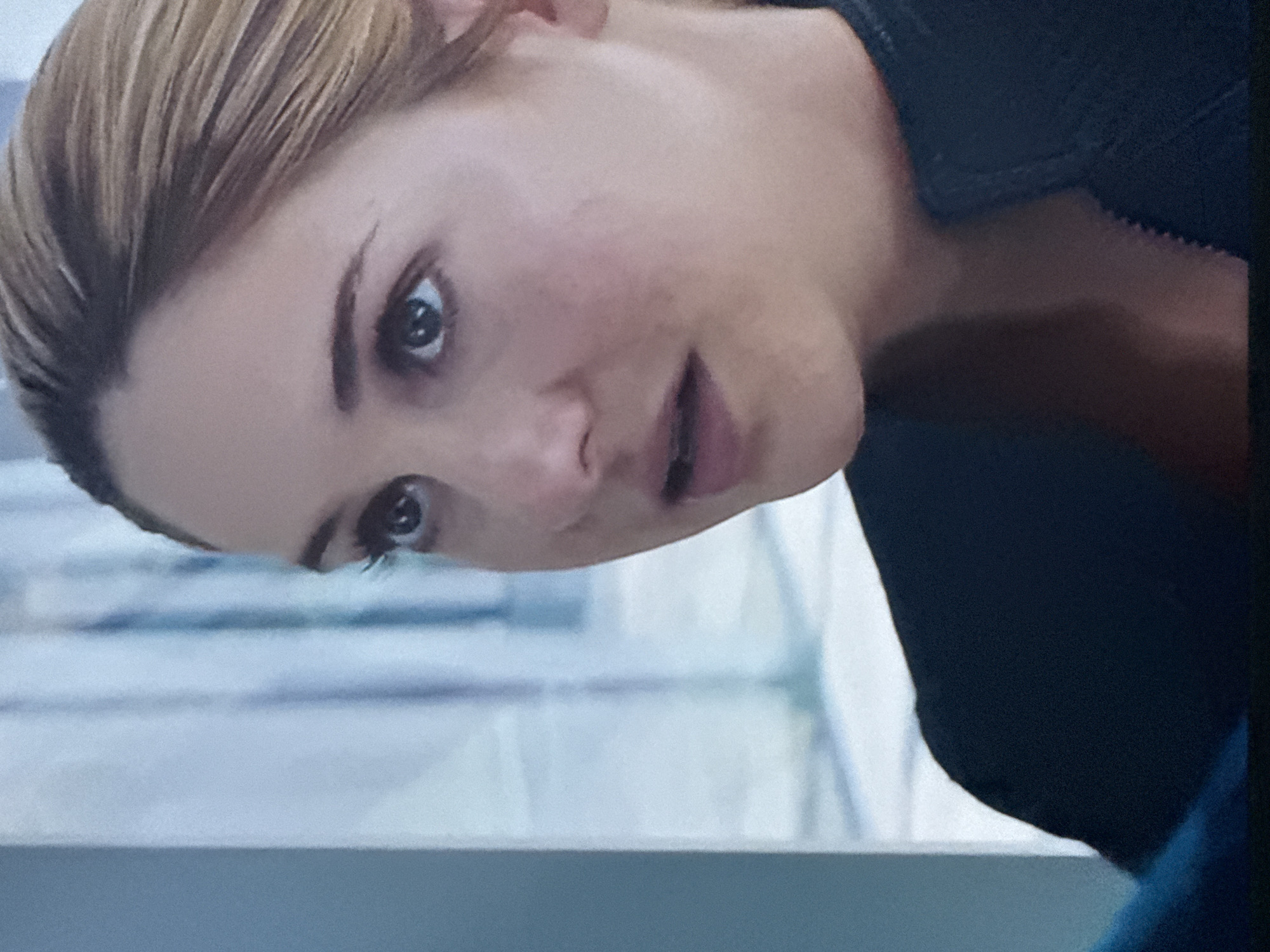 A close-up shot focuses on a fair-skinned woman with light brown hair. Her head is tilted, and her gaze is directed slightly downward. She has blue eyes, light eyebrows, and pink lips. She is wearing a dark-colored top, possibly black, with some textured or ribbed detailing visible near the collar. The background appears to be a blurred interior space with light, horizontal lines, possibly windows or reflective surfaces. The scene is not explicitly located in Mytishchi, Russia; there is no information to support this.