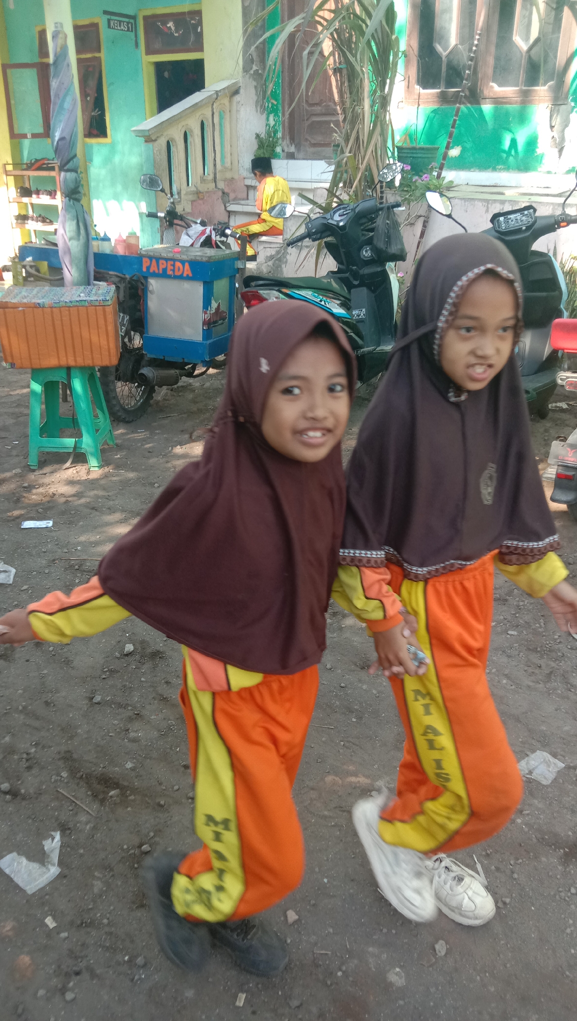 In Karang Anyar, Indonesia, two young girls wearing hijabs and matching orange and yellow school uniforms are captured in motion, seemingly playing. The girl on the left, with her head tilted and a wide smile, is looking towards the camera, her right arm extended slightly. The girl on the right is also looking slightly to the side and appears to be in mid-stride or jump, her right hand holding something small and metallic. They are standing on a dirt and gravel ground in front of a building with turquoise and cream-colored walls, possibly a school, indicated by a sign reading "KELAS 1" (Class 1). A blue food cart with "PAPEDA" written on it is visible behind them, along with a parked motorcycle. The environment suggests an outdoor, possibly urban or semi-urban setting on a sunny day.