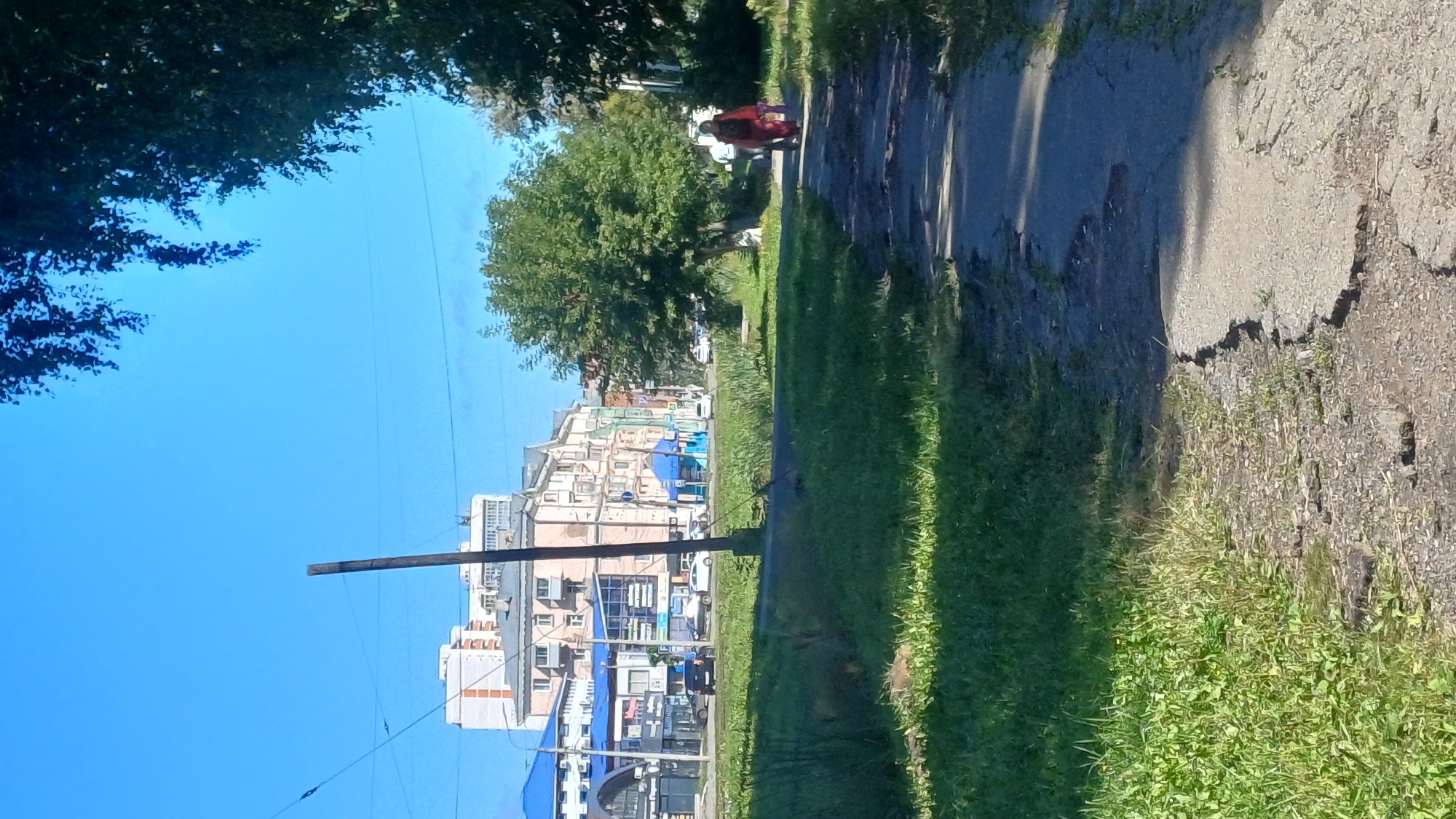 The image depicts a paved walkway in a city setting under a clear blue sky. The walkway is cracked and appears worn. On the right side of the walkway, a woman wearing a red garment is walking away from the camera, carrying a bag. To the left of the walkway, a grassy area is visible. In the background, there are several buildings, including apartment complexes and commercial establishments with blue awnings and signage. Cars are parked and driving on a street beyond the grassy area. A tall wooden pole stands near the center of the image, and trees with green foliage are present on the right and in the background. The overall scene suggests an urban environment during daylight hours.