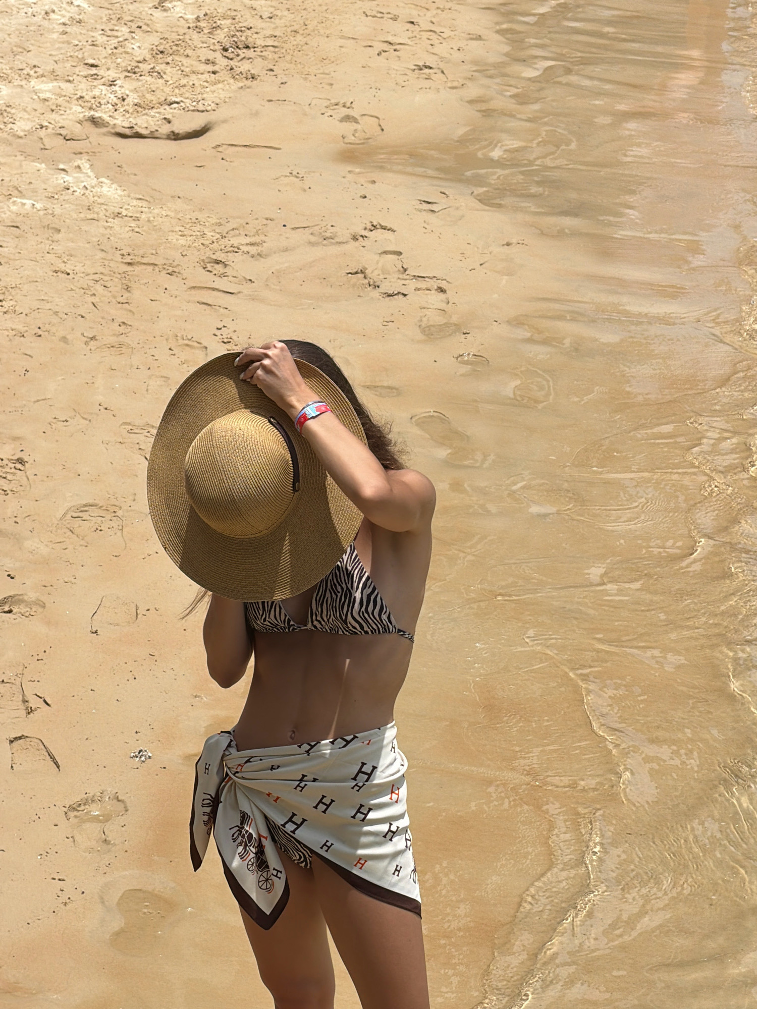A woman stands on a sandy beach, near the water's edge. She is wearing a zebra-print bikini top and a white sarong with a pattern of "H"s and horse-drawn carriages. She is holding a wide-brimmed straw hat over her face with her right hand, obscuring her features. A red and white wristband is visible on her left wrist. The sand is a light tan color with some small indentations. The water is shallow and clear, with ripples and reflections on the surface. The scene appears to be a sunny day at the beach. The image does not provide any specific information about the location being the city of Nha Trang, Vietnam.