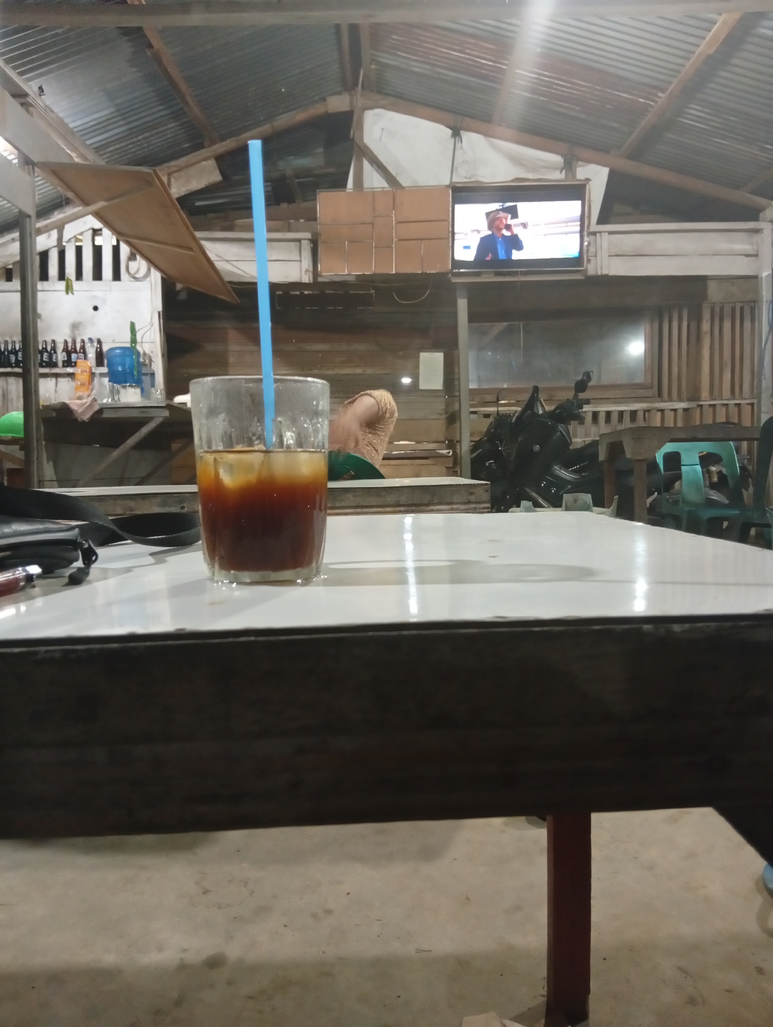 The image depicts the interior of a casual, open-air establishment in Lhokseumawe, Indonesia, likely a roadside eatery or warung. The scene is dimly lit, suggesting evening or night hours. In the foreground, a glass of iced coffee with a blue straw rests on a white-topped table. The table appears to be constructed from wood with dark metal legs.
In the background, a television screen mounted on the wall displays a person wearing a cowboy hat and blue shirt, possibly watching a program or movie. The structure of the establishment is visible, with a corrugated metal roof supported by wooden beams and rafters. Various objects are scattered throughout the background, including shelves with bottles, a water dispenser, what appears to be a motorcycle, and green plastic chairs. The flooring is a concrete surface. The overall impression is one of a simple, communal space designed for casual dining and entertainment.