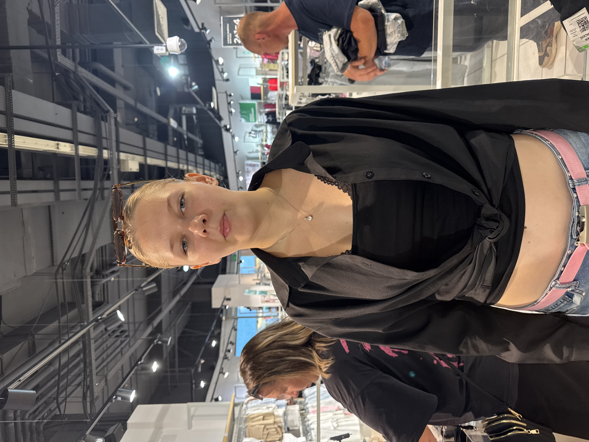 A young blonde woman stands facing the camera in a retail store. She is wearing a black collared shirt tied at the waist, revealing a black tank top and her midriff. She has on blue jeans with a pink belt and sunglasses perched on her head. Behind her, to the left, a woman with brown hair is browsing clothing racks. To the right, a bald man in a dark t-shirt is holding a stack of clothes. The environment appears to be a clothing store with racks of apparel, display shelves, and overhead lighting. The location is identified as a city in Perm, Russia.