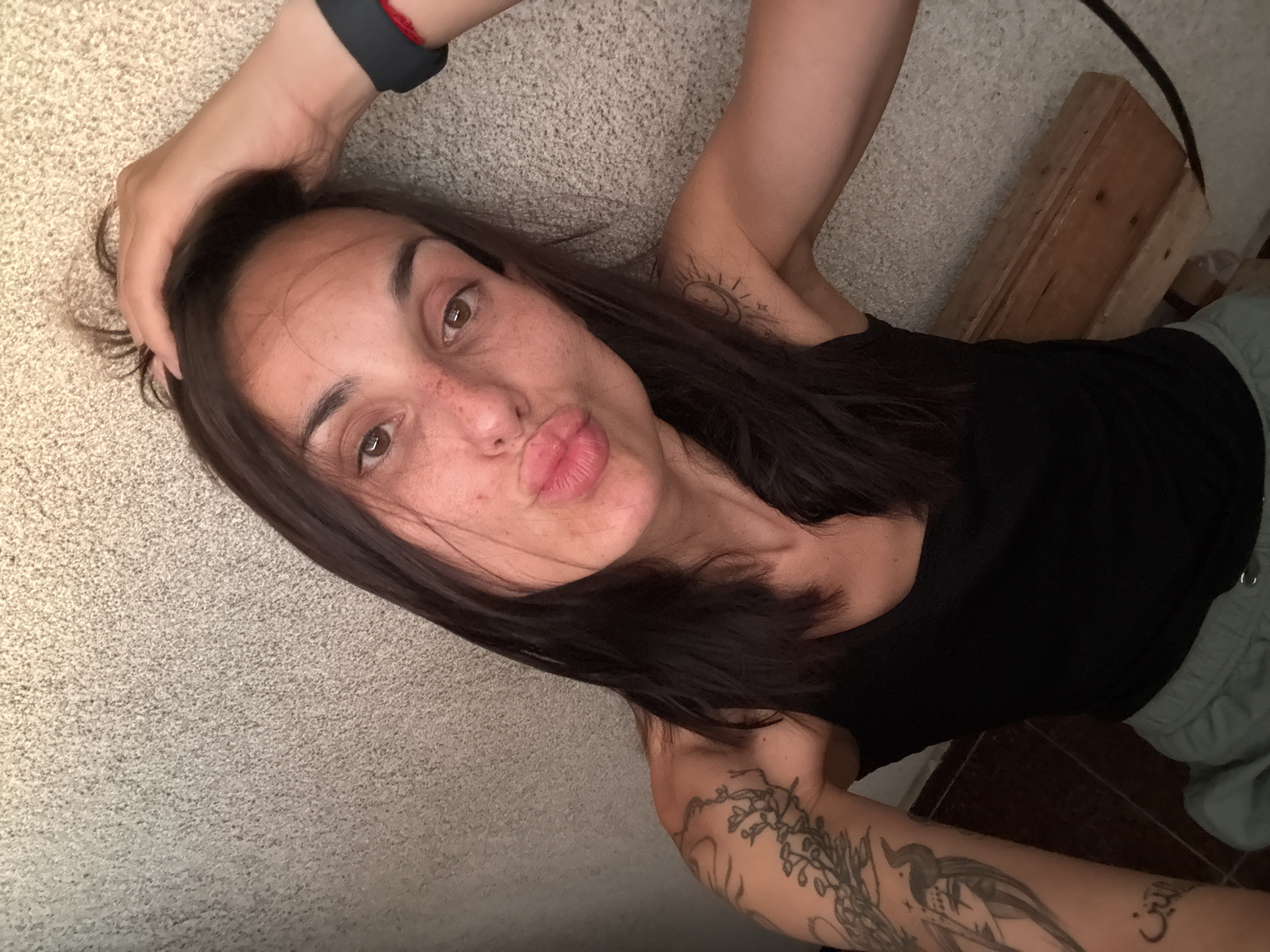 A woman with dark hair and freckles is looking at the camera and puckering her lips, her right hand is resting on her head. She is wearing a black tank top and grey shorts. She has tattoos on her left arm and a sun tattoo on her right shoulder. The background is a textured wall. The lighting appears to be natural, possibly from a window.