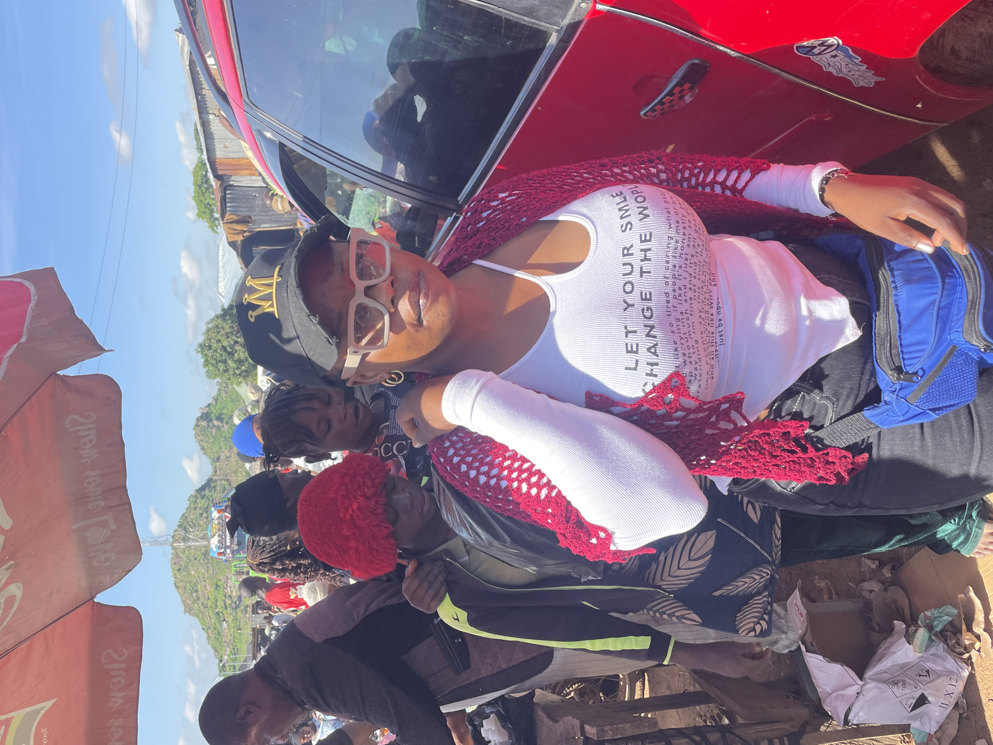 A medium shot captures a group of people outdoors in what appears to be a marketplace in Gura Top, Nigeria. In the foreground, a Black woman wearing a white tank top with a cutout neckline, a red crocheted shawl, dark pants, and a black baseball cap with a gold logo is looking towards the camera. She is also wearing large square-framed sunglasses. Next to her, a younger Black person with a red fuzzy hat is visible from the chest up. In the background, other people are milling about, and a red vehicle is parked to the right. The environment suggests a bustling outdoor market with stalls or shops in the distance. The lighting indicates it is daytime.