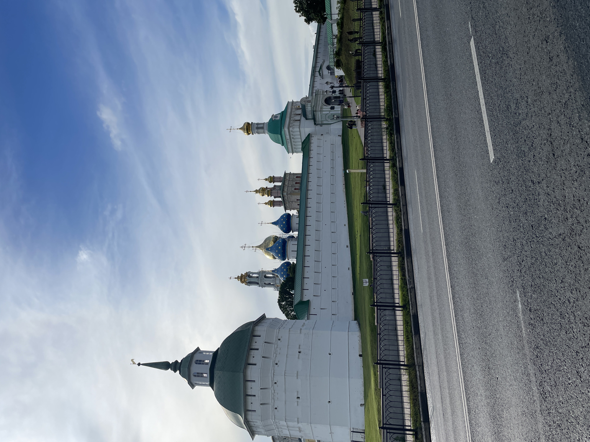 The image shows a view of the Trinity Lavra of St. Sergius in Sergiyev Posad, Russia. The foreground features a paved road with white lane markings and a black metal fence. Behind the fence is a well-maintained green lawn. The main subject is the complex of the Trinity Lavra of St. Sergius, a large white walled fortress with several towers and domes. The largest tower on the left is round and white with a green conical roof and a small flag on top. Further to the right, the white wall continues, punctuated by smaller towers and several domed churches. These churches have gold and blue domes, some topped with golden crosses. The sky above is a mixture of blue and white clouds. A few individuals are visible walking near the entrance of the complex and along the pathways within the grounds. The architecture suggests a historical and religious site.