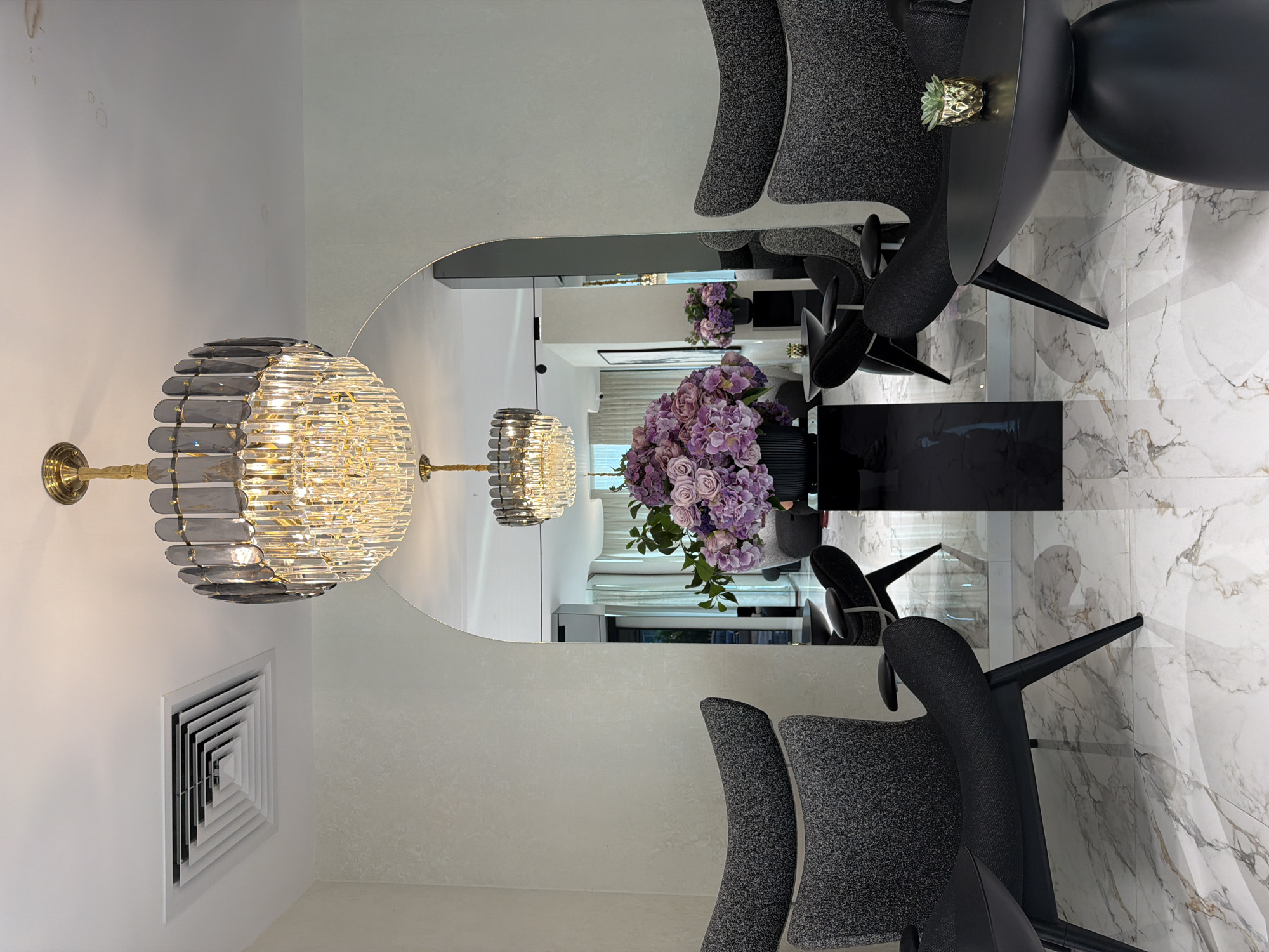 The image captures an interior setting, likely a waiting area or salon, featuring a large, round, mirrored wall. A crystal chandelier with a gold base is mounted on the wall. Seated chairs with gray, speckled fabric upholstery and curved black backs are positioned around a black, round table. On the table is a small decorative golden pineapple. A vase with purple flowers sits on a black stand, which is placed in front of the mirror. The mirror reflects the interior, creating an illusion of depth and repeating the elements within the room, including the chandelier and flowers. The floor is tiled with a white marble-like pattern. An air vent is visible on the wall. The image may have been taken in the city of Blagoevgrad, Bulgaria, but there is no specific evidence in the image to confirm this.