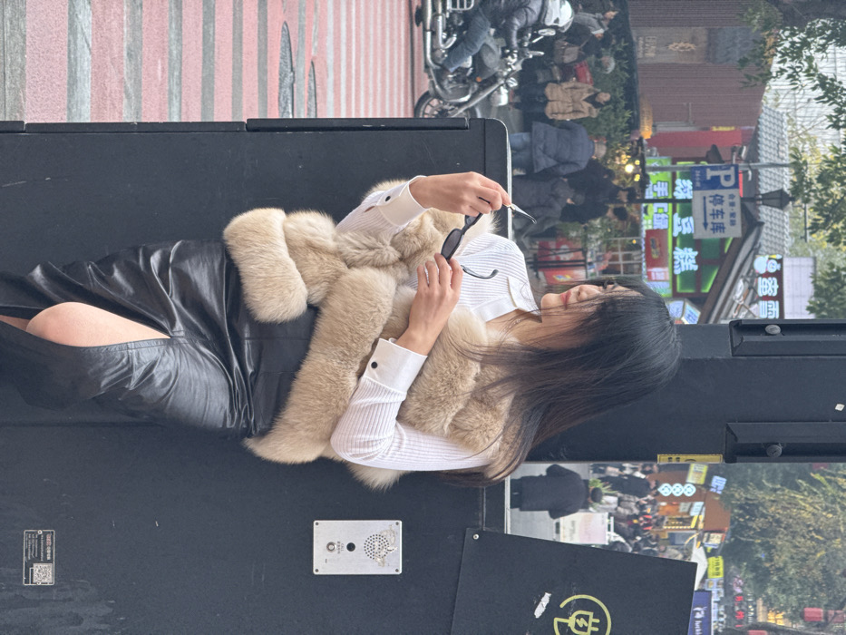 A woman sits on a dark surface, wearing a black leather skirt, a white ribbed top, and a beige fur scarf. She holds sunglasses in her hands. Behind her, a street scene unfolds with people walking past shops with lit signs. A motorcycle is parked near the upper edge of the frame. The photo is taken from a slightly elevated, angled perspective.