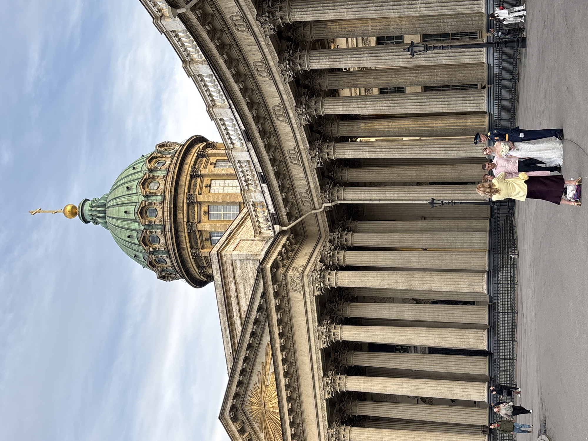 The image displays a grand architectural landmark, Kazan Cathedral, in Saint Petersburg, Russia, under a partly cloudy daytime sky. The building, a prominent example of Neoclassical architecture, occupies the majority of the frame. Its most striking features include a large, ribbed green dome topped with a golden finial and a cross, and an expansive colonnade of tall, fluted columns with ornate Corinthian capitals. Decorative elements, including what appear to be crowns or stylized symbols, adorn the entablature above the columns. A golden sunburst motif is visible on a pediment towards the lower left.
In the mid-ground, a celebratory event is taking place. A wedding couple stands prominently: the bride in a white gown holding a white bouquet, and the groom in a dark blue military-style uniform with a cap. Nearby, a woman in a yellow top and dark skirt, possibly a photographer, is focused on the couple, while another man in a light pink shirt stands beside them. Other people are visible further back and to the sides, including a figure in a white suit and several individuals near the railings and columns. A black, ornate lamppost stands in the foreground, separating the people from the cathedral's massive base. The scene captures a vibrant wedding photoshoot against the historic backdrop of the iconic cathedral. No discernible text is visible.