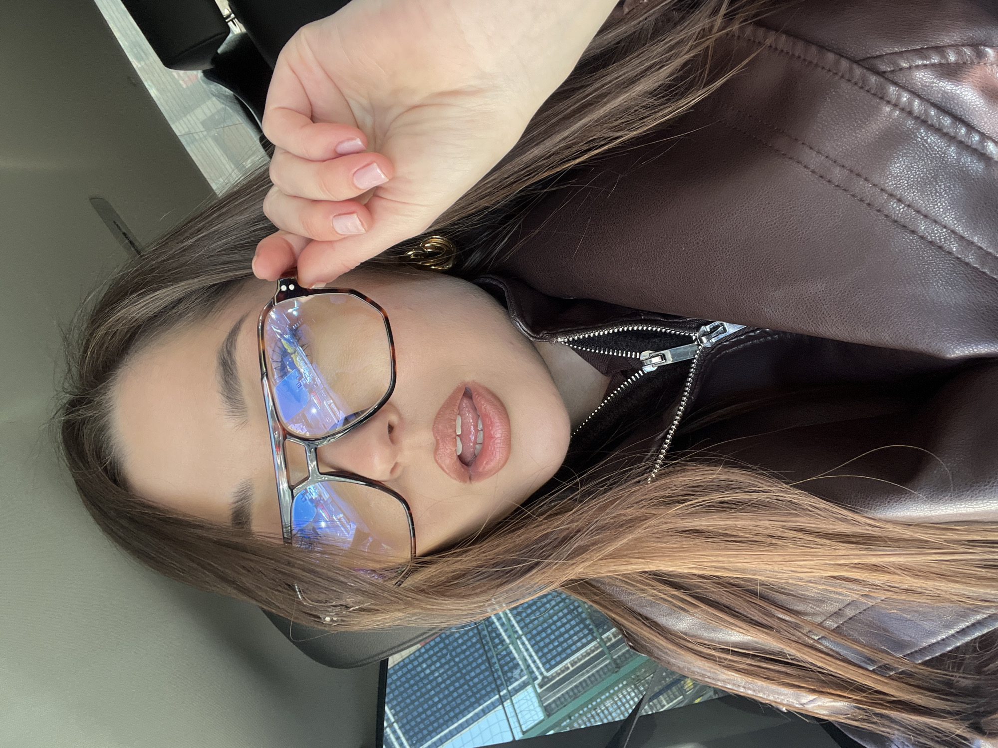 A young woman is seen in a close-up selfie, holding her glasses at an angle. She has long brown hair and is wearing a brown leather jacket with a visible zipper. Her lips are slightly parted. Reflections of buildings are visible in her glasses, indicating an urban environment. The location is identified as Khimki, Russia, as visible in the reflections in her glasses.