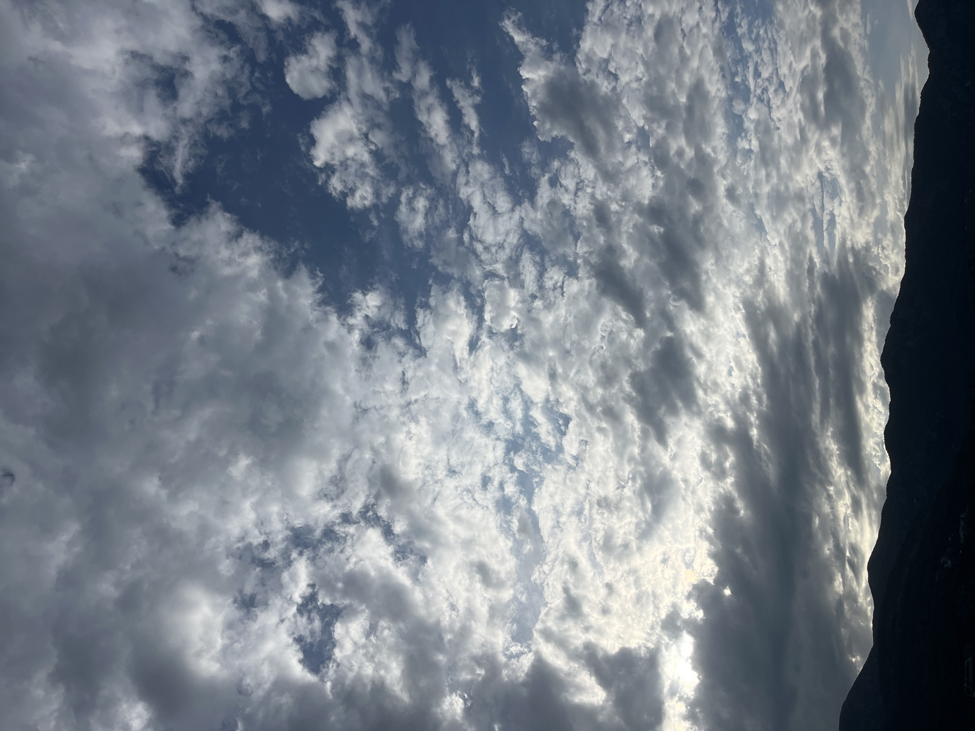 The image depicts a sky filled with numerous small, puffy clouds, with patches of blue sky visible in between. The clouds range in color from bright white where illuminated by the sun, to shades of gray. The silhouette of a dark, jagged mountain ridge is visible on the right side of the frame. The location is Budva, Montenegro.
