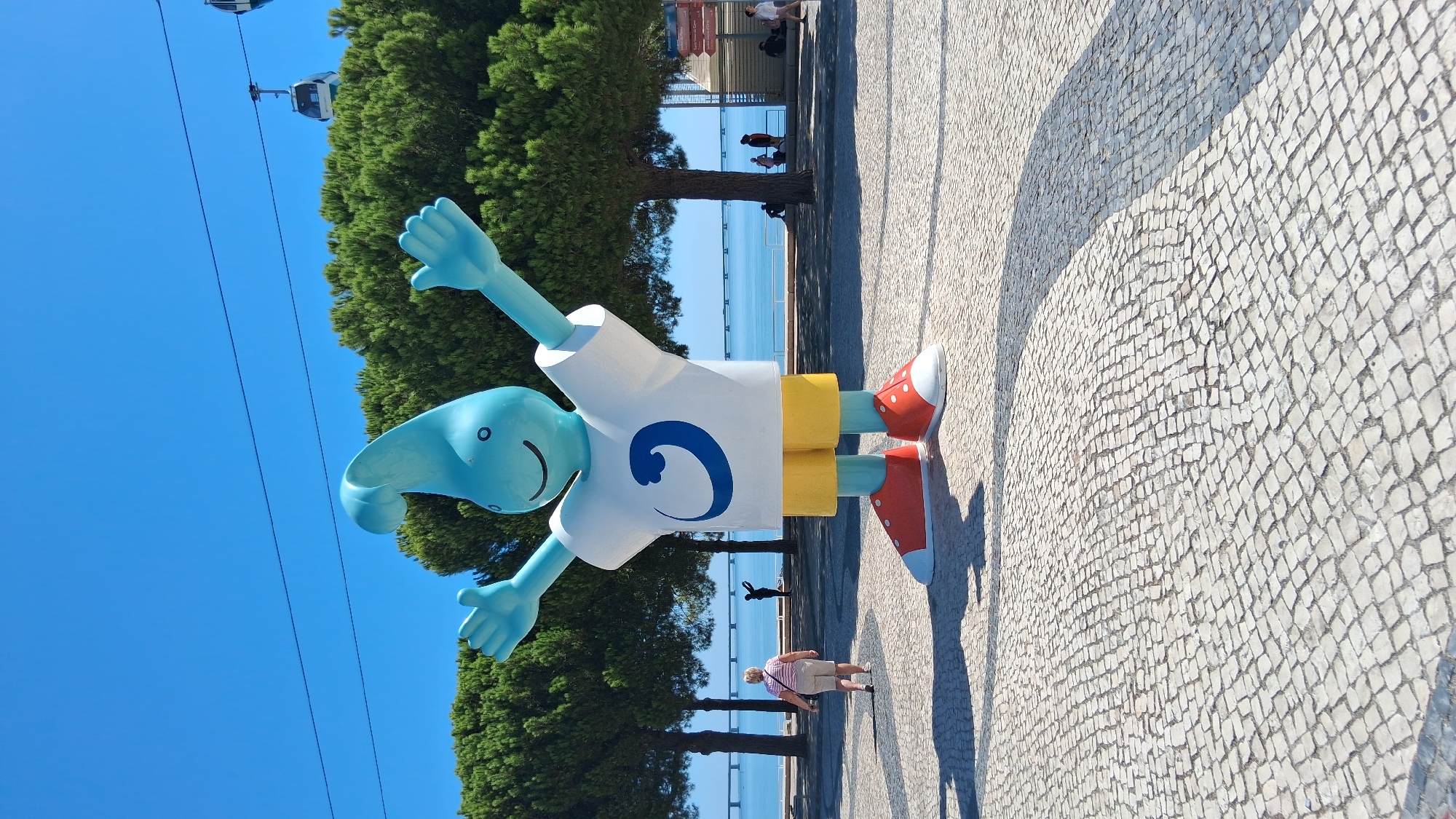 This image captures a large, blue, cartoonish mascot with its arms outstretched, standing on a cobblestone plaza. The mascot is wearing a white t-shirt with a blue wave logo, yellow shorts, and red and white sneakers. Behind the mascot, a woman walks away from the camera. Further in the background, a bridge spans a body of water, and cable cars traverse the sky. Trees line the waterfront, and people are visible in the distance. The scene suggests a sunny day with clear blue skies. This location is the Parque das Nações in Lisbon, Portugal, a district developed for Expo '98.
