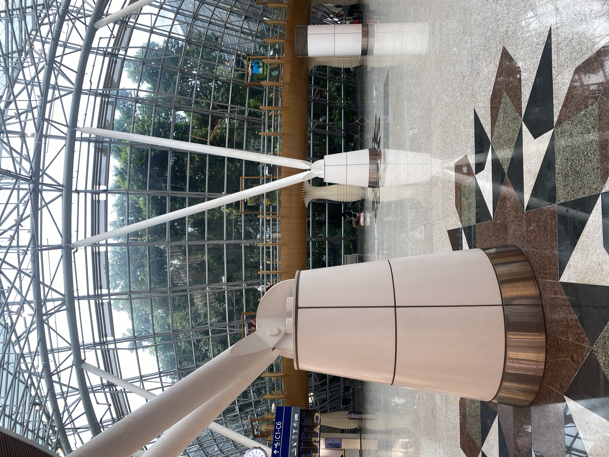 The image depicts the interior of a modern airport terminal in Sepang, Malaysia. The scene is dominated by a large, geodesic dome-like glass and metal roof structure, allowing natural light to illuminate the spacious interior. Large, white, conical pillars support the roof. In the foreground, a section of the floor is visible, characterized by an intricate geometric pattern of polished granite tiles in shades of brown, black, and green.
In the middle ground, a raised walkway with wooden railings is visible, with people walking along it. Various amenities, including seating areas and cylindrical structures, are scattered throughout the terminal. In the left of the image, a blue directional sign indicates "↑C1-C6" and displays various icons. A clock is also visible on a pillar to the left. In the background, lush green trees are visible through the glass ceiling, suggesting an outdoor environment beyond the terminal.
Several individuals are present, mostly in the background. On the raised walkway, a person is walking. In the lower left, two individuals are seated. To the right, a person is standing near a trolley. The overall atmosphere is one of a clean, well-lit, and organized public space, characteristic of an international airport.