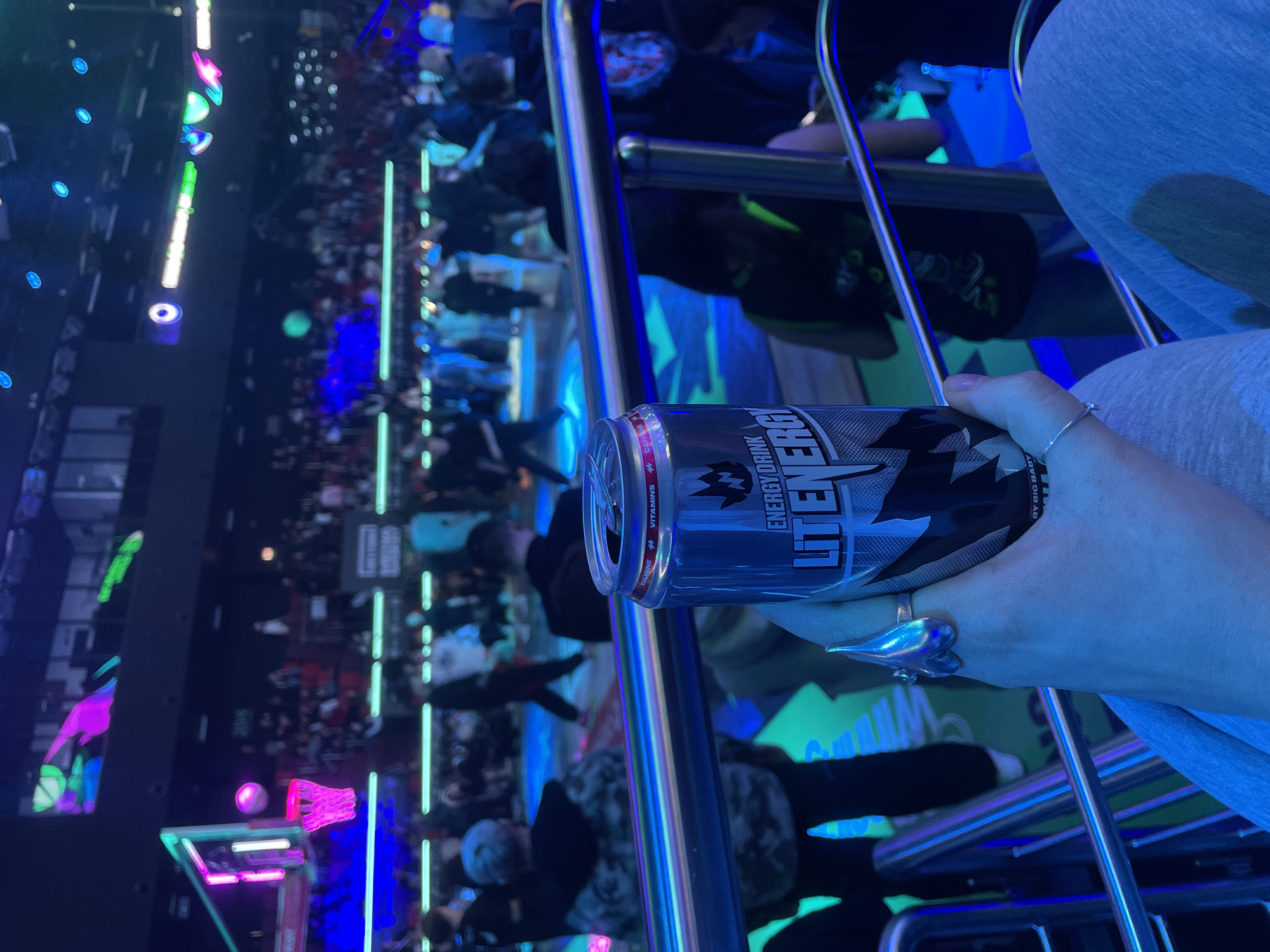 A person is holding a can of "LIT ENERGY" drink. They are seated in a chair with chrome armrests. The background shows a crowded indoor space with bright neon lighting, including pink and blue hues. The location appears to be a gaming or entertainment venue. There's a basketball game or activity visible in the background. The image was likely taken indoors. It is unknown if the location is in Moscow, Russia.