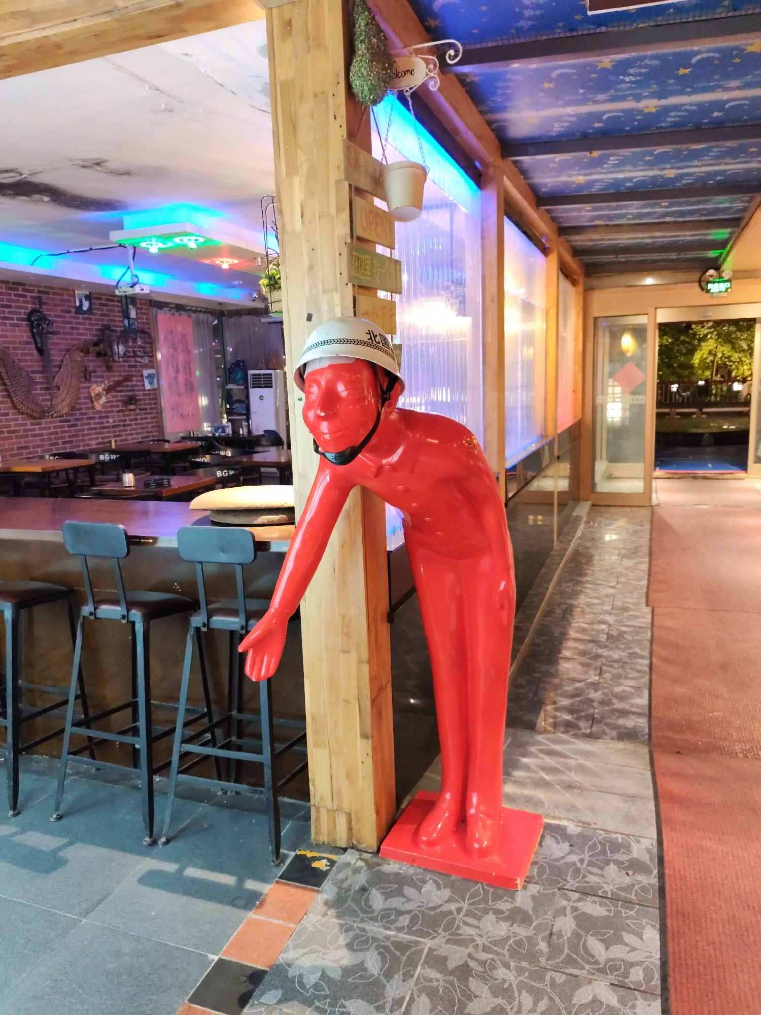 The image depicts the interior of a restaurant or bar in Lindian Town, China. In the foreground, a bright red, human-shaped mannequin wearing a white helmet is positioned leaning against a wooden pillar. The mannequin's right arm is extended downwards, with its hand open. Behind the mannequin, a bar with several black stools is visible. The background shows a brick wall with decorations, tables and chairs, and brightly colored LED lighting on the ceiling. The flooring consists of stone tiles. Through a glass partition and doors in the background, an outdoor area with trees and possibly a pool can be glimpsed. The overall impression is of a lively and perhaps unconventional establishment.