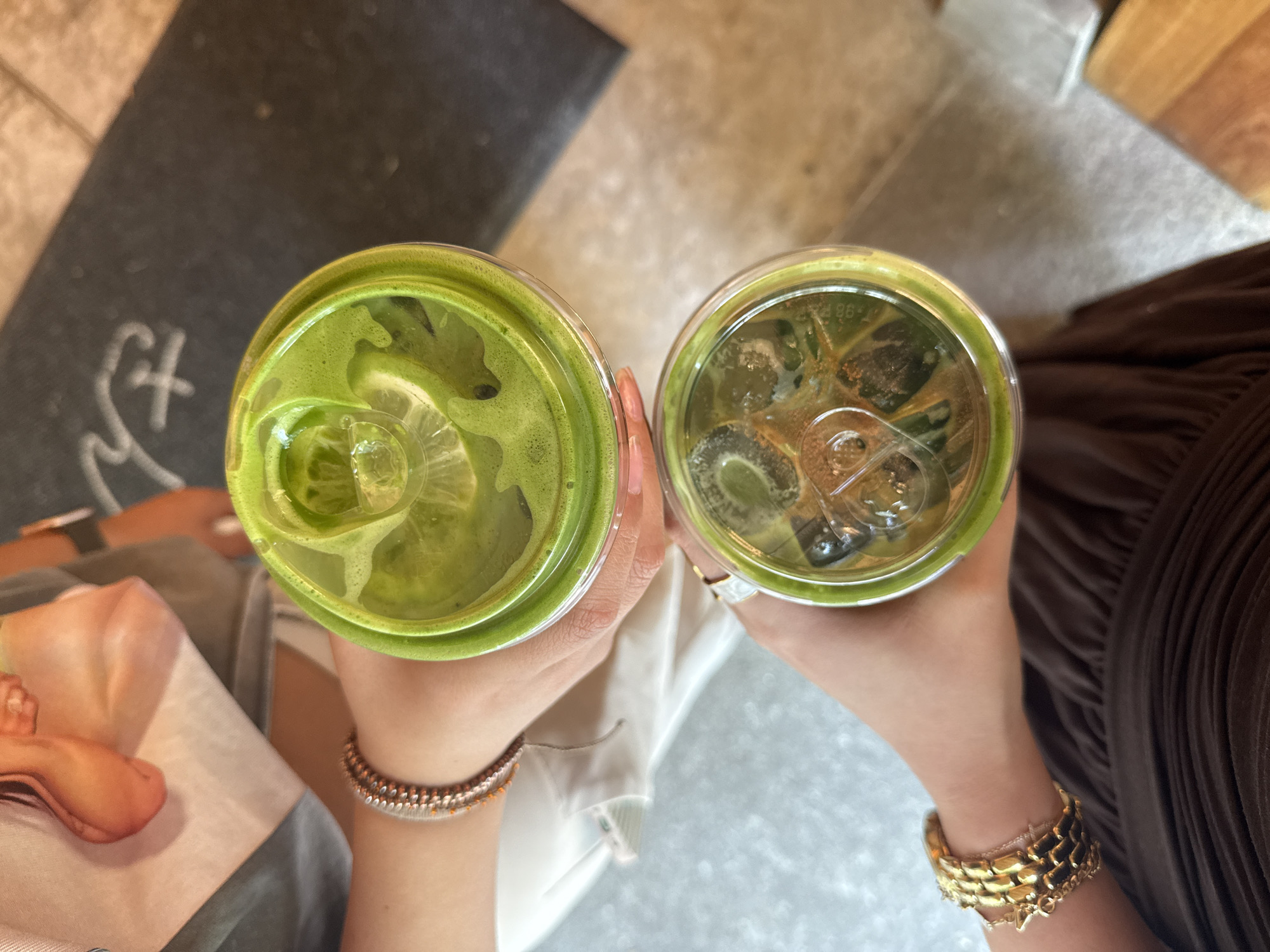 An aerial perspective showcases two individuals holding what appear to be beverages in plastic cups, potentially matcha drinks, set against a backdrop of a street in Mexico City, Mexico. The person on the left wears a light-colored shirt and a beaded bracelet, while the one on the right is dressed in brown and sports a gold bracelet. The beverages contain ice and feature a green hue.