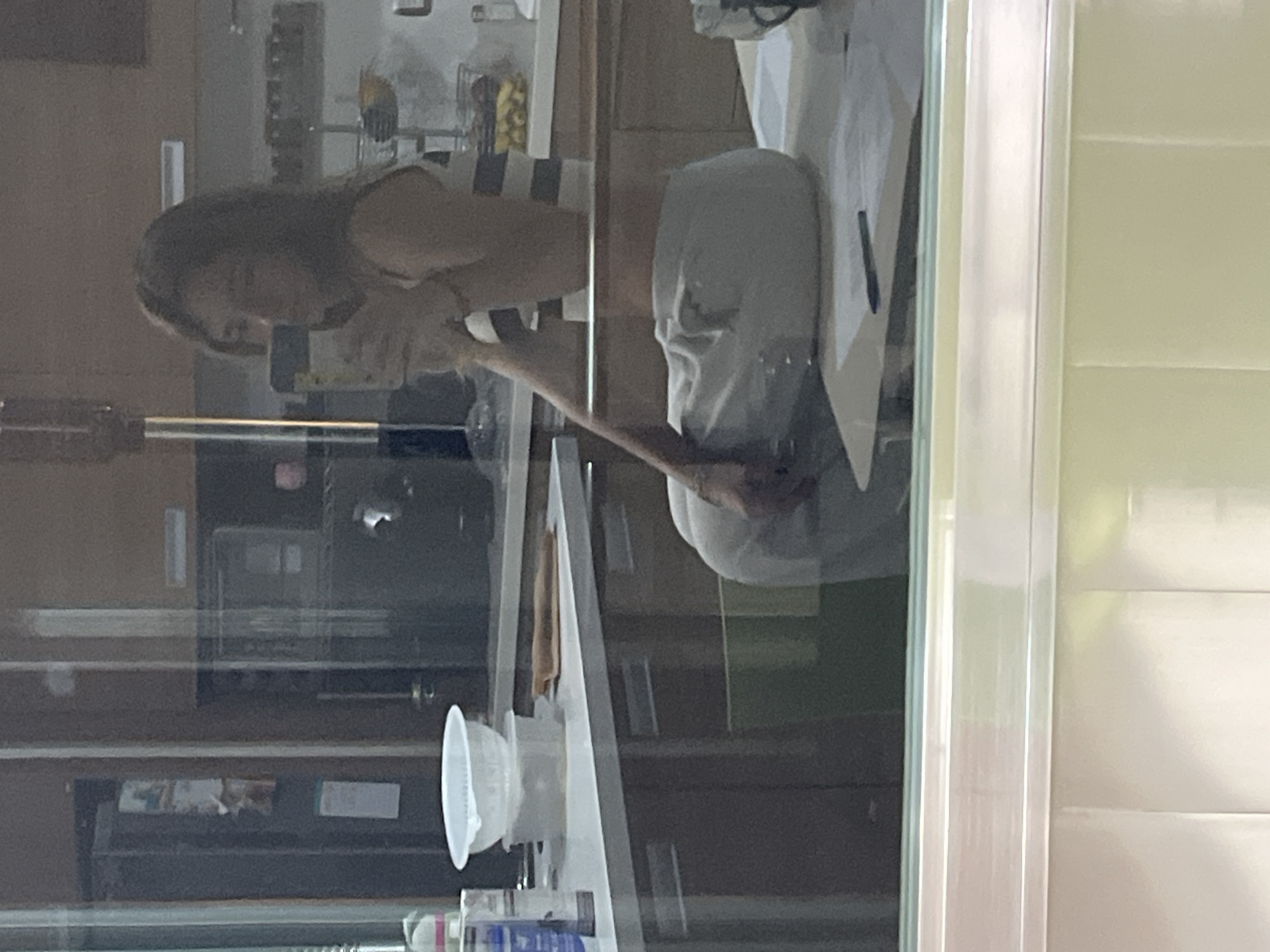 A reflection of a woman is seen in a window. The woman is wearing a striped top and gray pants. She is holding something to her mouth with her right hand. In the foreground, there is a kitchen counter with a white bowl and various bottles of cleaning supplies. The location is possibly San Bernardino Tlaxcalancingo, Mexico.