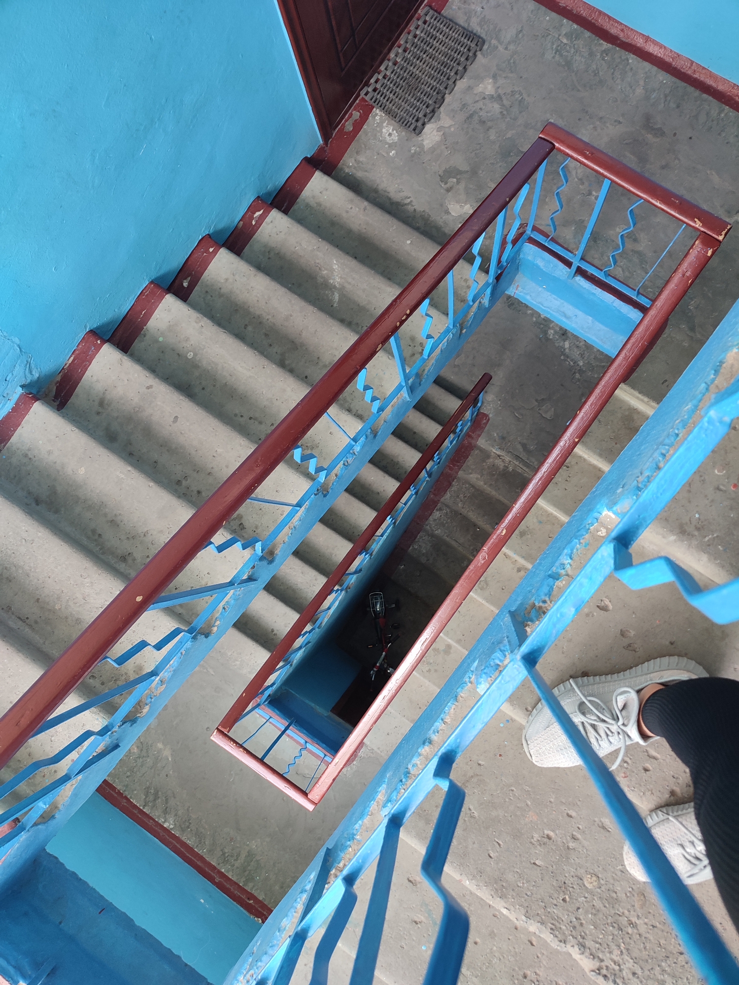 A first-person perspective shot from above shows a person's feet and lower legs on a concrete staircase with blue railings and a dark brown handrail in Yaresky, Ukraine. The person is wearing light gray athletic shoes and black pants. The staircase, which appears to be in a public building like an apartment complex or an office building, is utilitarian with worn concrete steps. The blue railings have a zig-zag pattern and show signs of wear and tear, with some chipping and debris. The walls are painted a vibrant blue. The image captures the descent down the stairwell, with multiple landings visible. The overall impression is one of a functional, somewhat aged, but still accessible urban environment.