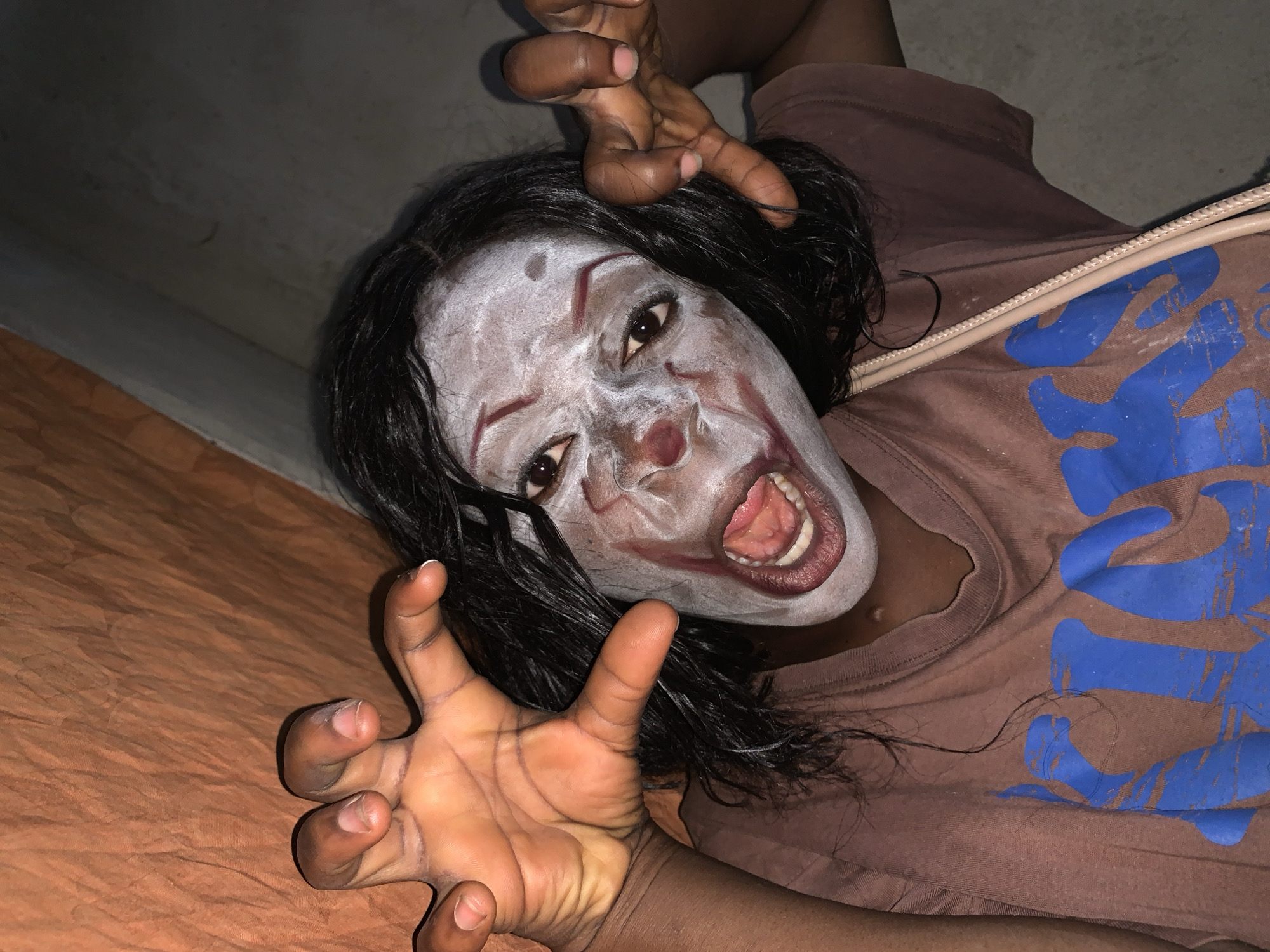 A person with dark skin and black hair is lying down with their face angled toward the camera. The person is wearing white clown makeup with red accents around the eyes and mouth. The person's mouth is open and their hands are positioned near the camera with fingers outstretched. The person is wearing a brown t-shirt with blue lettering and a zipper pull. The background is dark and indistinct, but there appears to be a brown-orange surface below the person and a dark wall above. There is no information to indicate this image was taken in Agban, Nigeria.