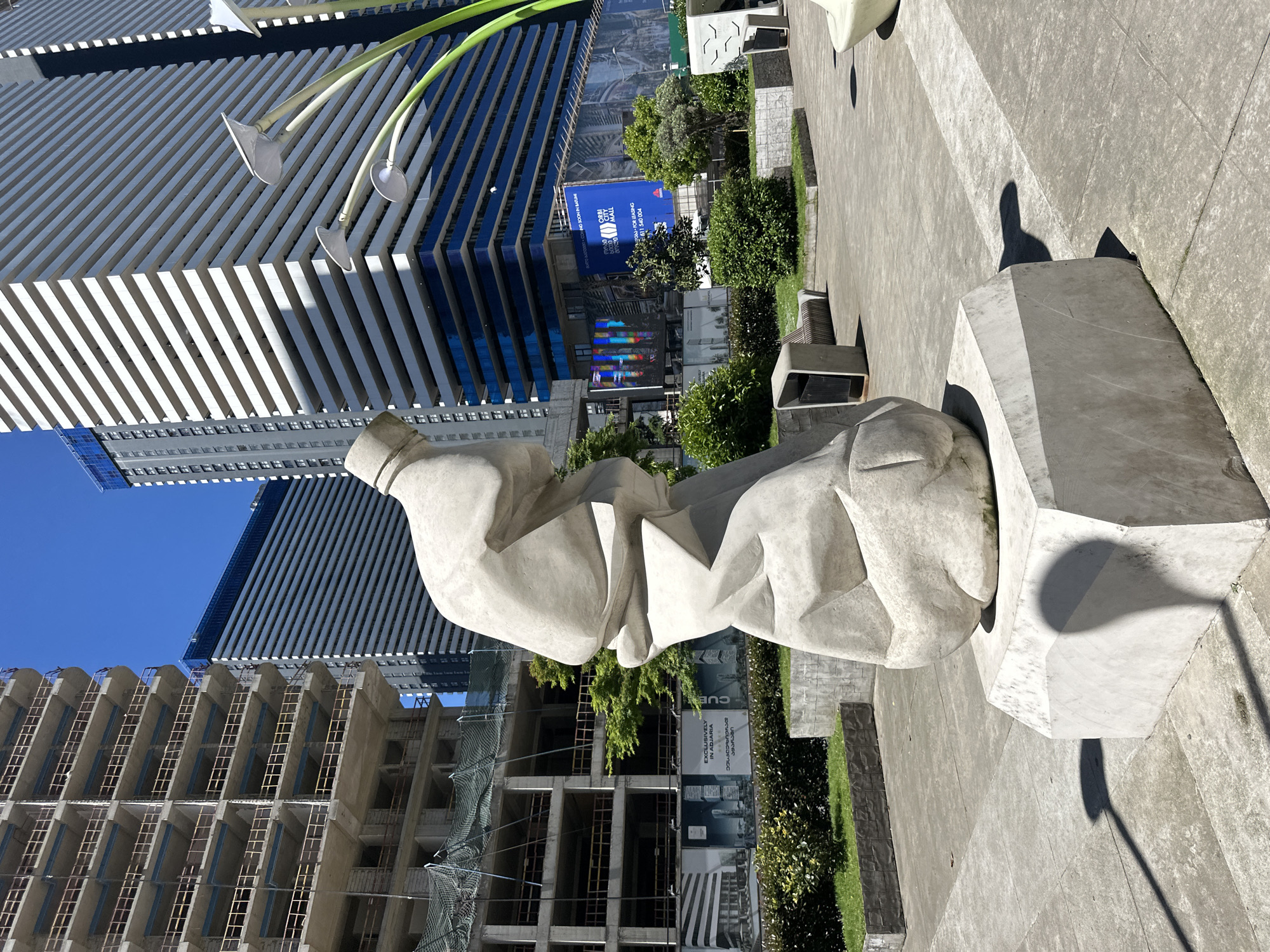 The image captures a contemporary outdoor setting in Batumi, Georgia, showcasing a large, abstract sculpture constructed from light-colored stone or concrete. The sculpture rests on a similarly textured pedestal, situated on a paved plaza with integrated greenery and seating.
The environment includes modern architecture, with a partially constructed building featuring scaffolding and a completed structure with distinctive horizontal stripes and blue glazing. Advertising signage is present, indicating commercial leasing opportunities and mentioning "ORBI MALL."
The lighting suggests a sunny day. There are no people visible in the image. The scene is urban, exhibiting a mix of artistic installation and commercial development.