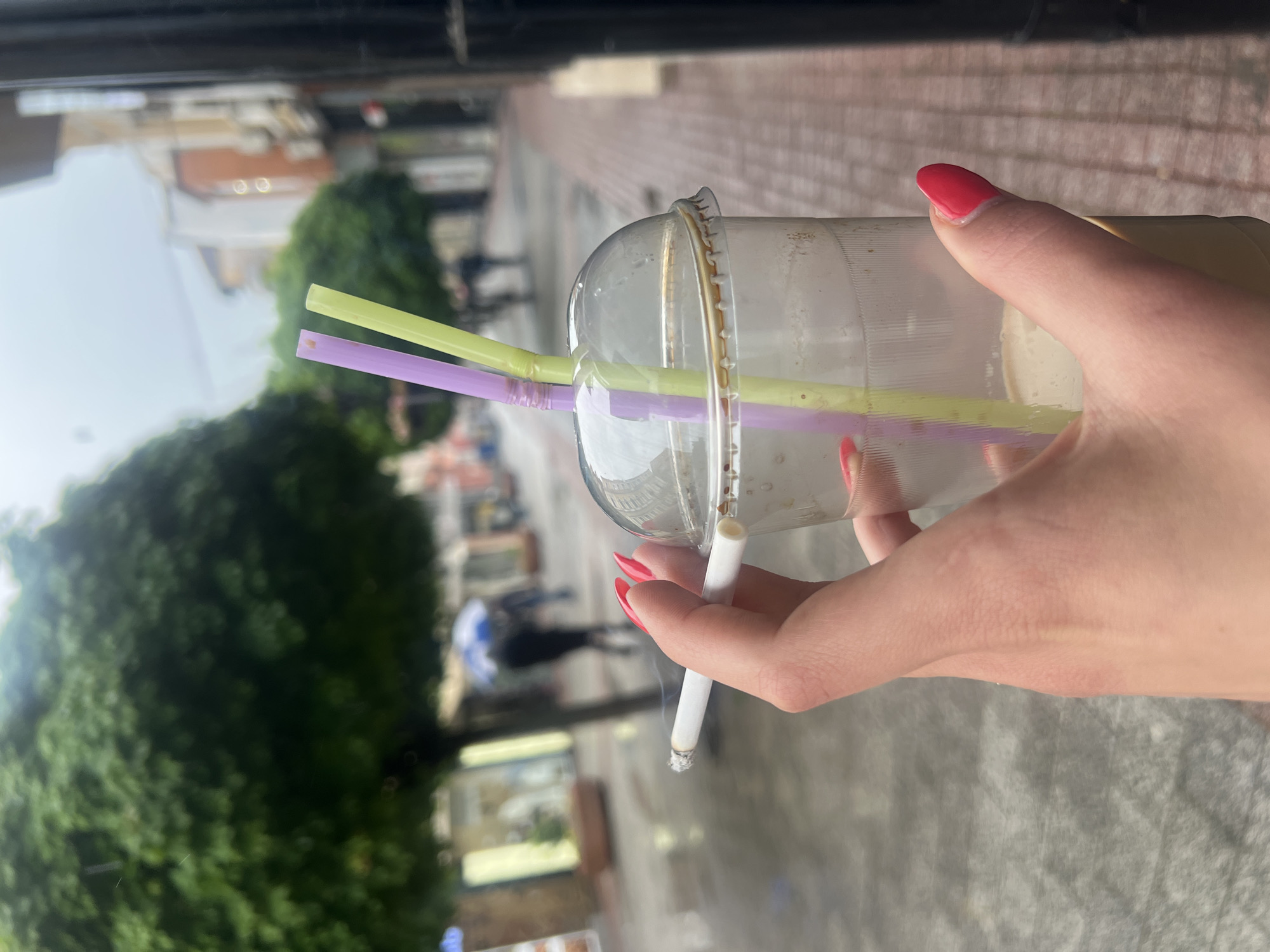 A person holds a disposable clear plastic cup with a domed lid, containing a light-colored liquid, in their right hand. Two plastic straws, one pale yellow and one lavender, are inserted through the lid. In the same hand, the person holds a lit cigarette. The person has red nail polish. The background is an out-of-focus urban scene featuring trees, buildings, and pedestrians. Based on the architecture and details visible, the location appears to be in Plovdiv, Bulgaria. The scene appears to be outdoors, possibly on a street or sidewalk.