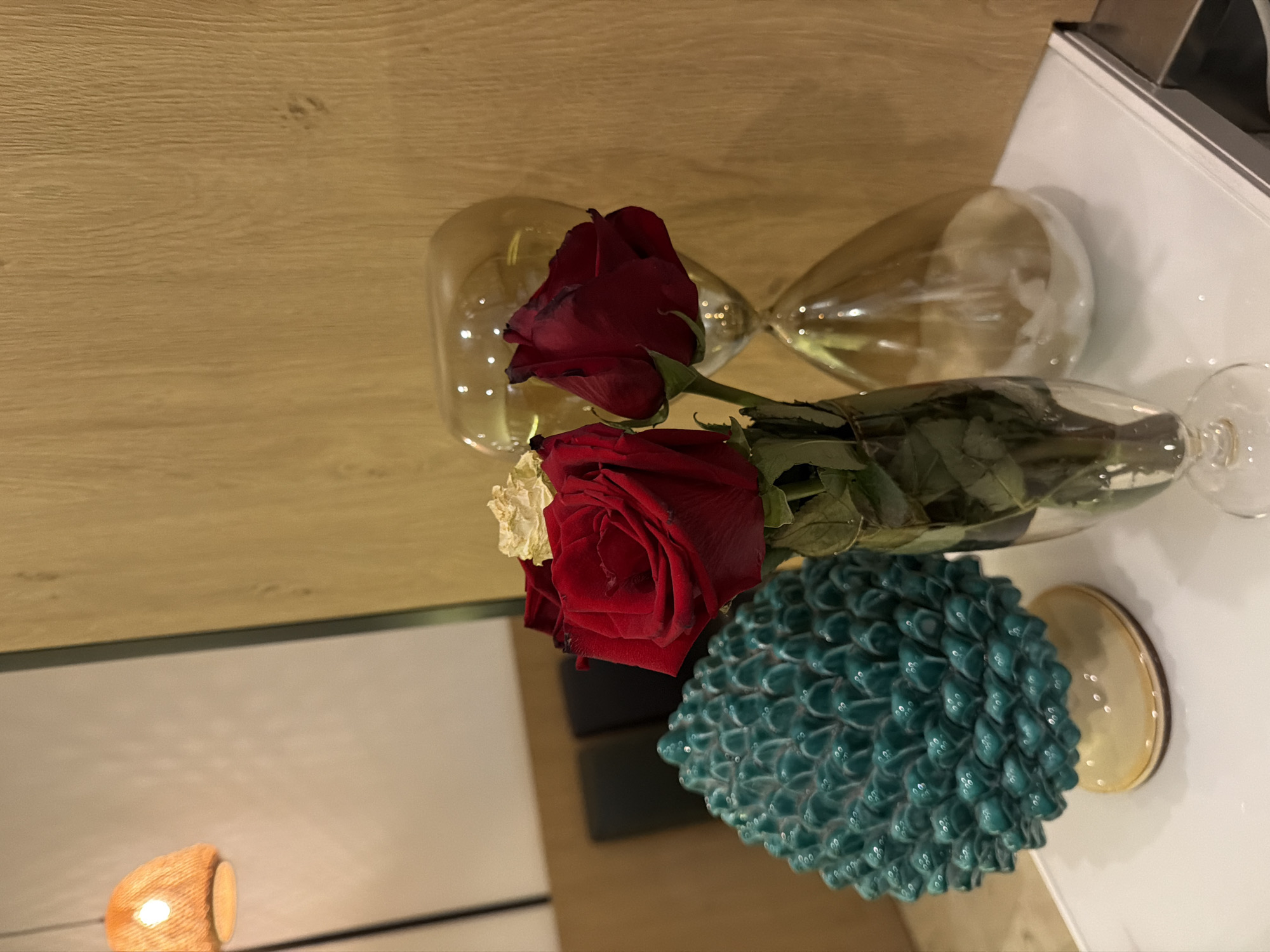 A still life arrangement sits on a white surface. In the foreground, a clear glass vase holds three red roses, one with a dried yellow petal. To the left of the roses is a turquoise ceramic object shaped like a pinecone, with a light-colored base. Behind the roses, a large, amber-colored glass hourglass stands on its side. The background consists of a light-colored wall on the left and a warm-toned, wood-paneled wall on the right. A woven lampshade is visible in the upper left corner. The overall scene appears to be indoors, possibly in Otranto, Italy, given the decorative ceramic style.