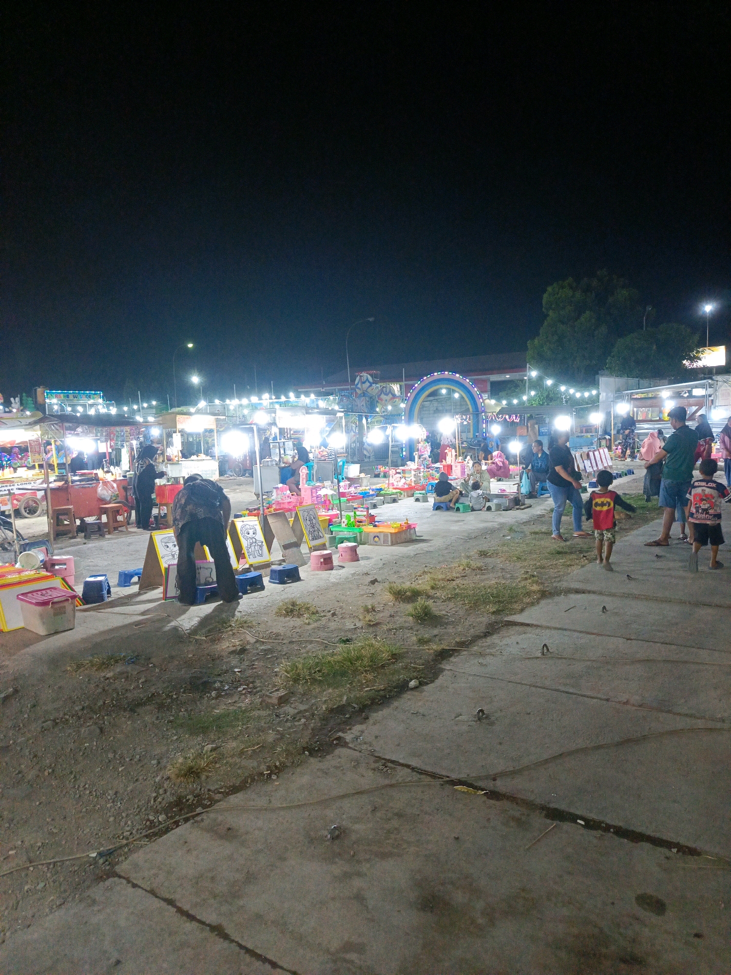 This is a night-time photograph of a busy outdoor market or carnival located in Sedati Agung, Indonesia. The scene is illuminated by numerous string lights and stall lighting, creating a vibrant and somewhat chaotic atmosphere.
In the foreground, a person is bent over, seemingly examining items laid out on the ground. Various plastic containers and stools are scattered around. A dirt path with patches of grass runs alongside a paved walkway.
The mid-ground features numerous market stalls, some appearing to sell food and others offering toys or games. Several people are visible browsing the stalls or interacting with vendors. Children are present, with two young boys prominently featured in the right side of the frame. One boy is wearing a red shirt with Batman imagery, and another is wearing a shirt with "ROBLOX" text.
In the background, a large, brightly lit archway, possibly part of a ride or entrance, is visible. More stalls and lights stretch into the distance. Trees and a utility pole are also discernible in the background, indicating an urban or semi-urban environment. The overall impression is one of lively evening commerce and entertainment.