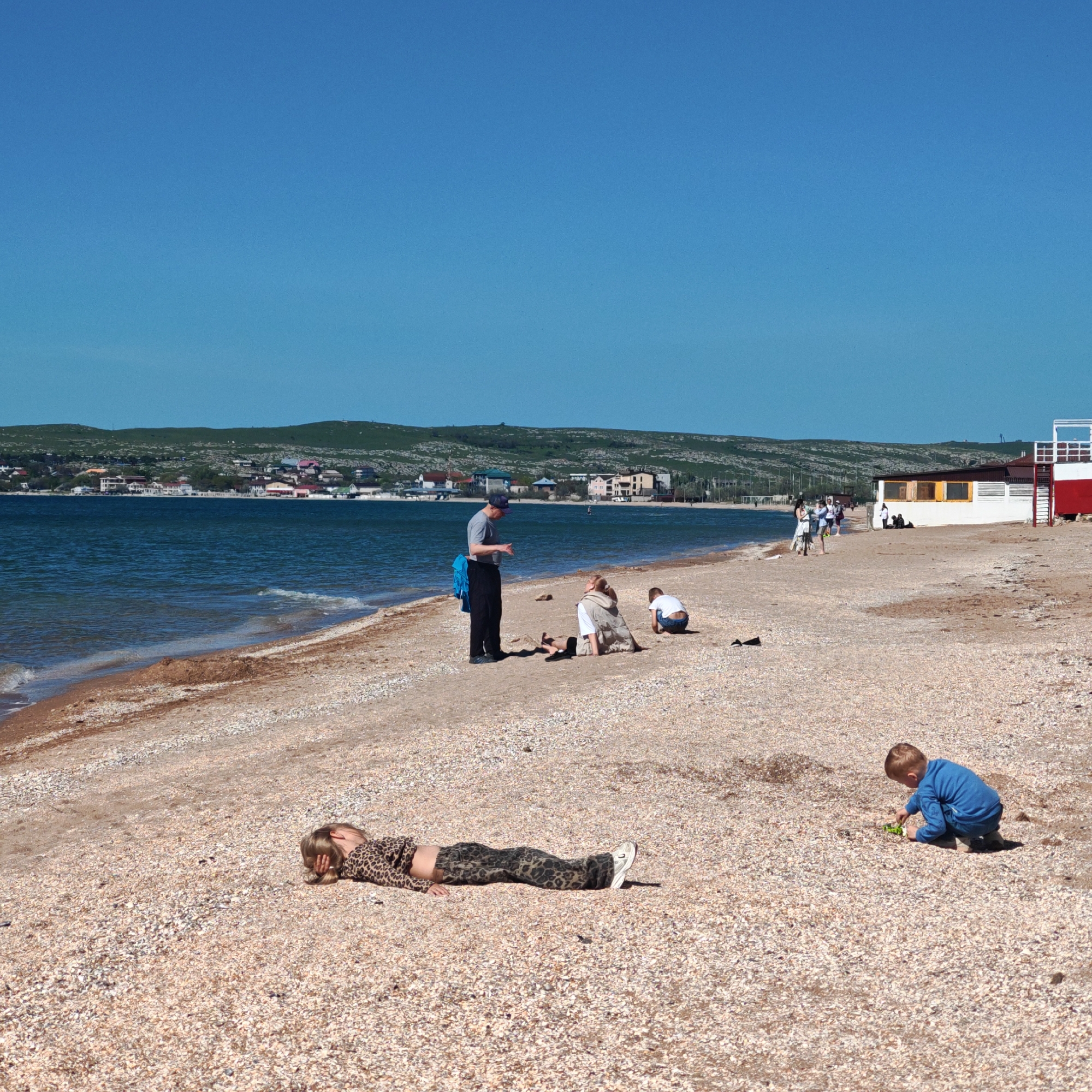 The scene is a beach in the city of городское поселение Щелкино, Ukraine. The sky is clear and blue. The beach is covered with shells. The water is dark blue and there are small waves. In the distance is a green hill with buildings. In the foreground, a girl is lying on her stomach on the shells. She is wearing a leopard print outfit. A boy is kneeling on the shells, playing with a green toy. Further back, a man is standing and talking to two children who are sitting on the ground. In the background, people are walking along the beach near a white and red building.