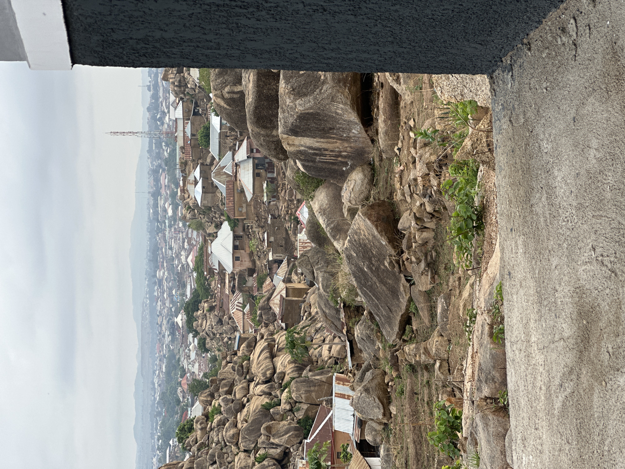 The image depicts a hillside view in Jos, Nigeria, characterized by numerous large, rounded rocks interspersed with dwellings and vegetation. The houses are mainly modest structures with corrugated metal roofs, clustered amidst and around the rocks. The hillside transitions into a more densely built urban area in the distance, partially obscured by haze. A communication tower is visible on the horizon. In the foreground, parts of a building structure, painted gray and black, frame the scene. The overall impression is one of a settlement uniquely integrated into a rocky and undulating landscape.