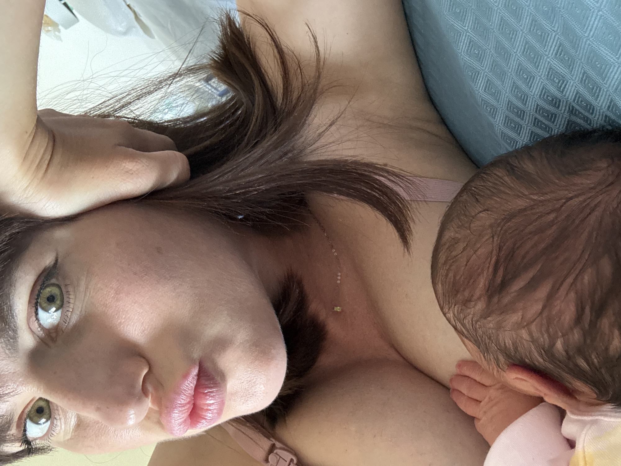 This media file captures a serene and intimate moment between a mother and her infant, likely situated in Aprilia, Italy. The image is taken from a slightly elevated, close-up perspective, with the mother lying down and engaging with the camera.
The primary subject is a woman, presumably the mother, whose head is tilted, revealing her striking green/hazel eyes, which gaze directly at the viewer. Her full, pink lips are slightly parted. She has medium-length brown hair that cascades around her face and shoulder. A delicate gold chain necklace with a small green charm adorns her neck and chest. Part of a light pink bra strap is visible on her shoulder. Her expression appears calm and tender.
Next to her, the back of an infant's head is visible, characterized by dark hair. The baby's tiny hand is seen resting near the mother's chest, suggesting it is either being held close or is in the act of breastfeeding. The baby is wearing a light-colored garment, possibly white or pale pink/yellow.
The setting appears to be indoors, likely a bedroom or living space. A light blue fabric with a textured, diamond-shaped pattern is visible, serving as a pillow or part of a bedspread. The lighting is soft and diffused, indicating daytime. No specific time of day beyond "daytime" is discernible, nor are there any explicit location cues within the image itself. There is no visible text present.