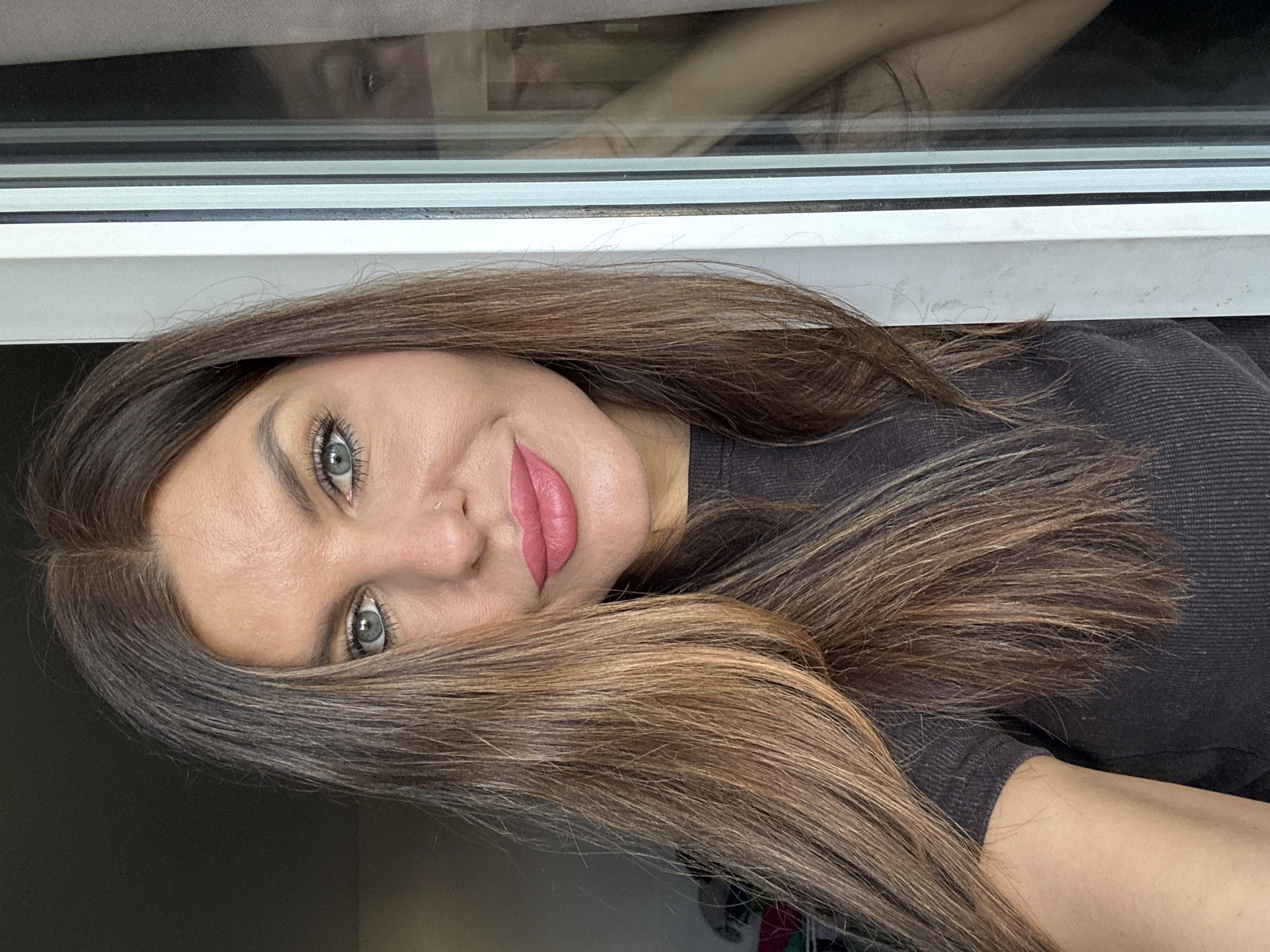 A medium close-up shot captures a woman with fair skin, light eyes, and long, wavy brown hair with lighter highlights. She is wearing a dark, possibly black, garment. The woman has a nose ring and is wearing pink lipstick. The photo appears to be taken indoors, near a window frame, with a reflection visible in the windowpane. There is no indication that this image was taken in the city of Chaikovsky, Russia.