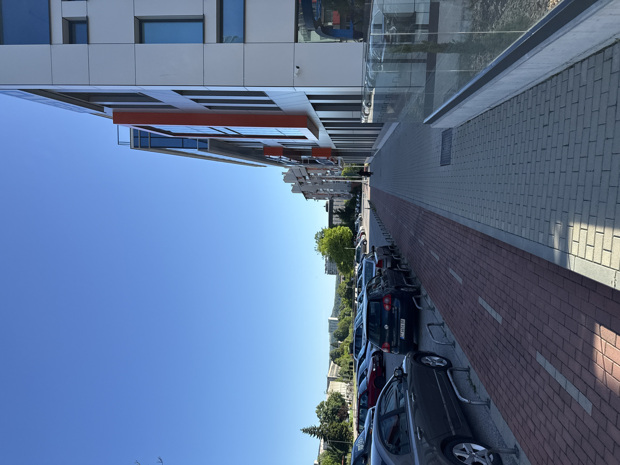 The image captures a street scene in Gabrovo, Bulgaria, with a modern building on the left side and a row of parked cars along the right. The building has a white and grey facade with orange accents and reflective windows. A narrow pedestrian pathway runs alongside the building, paved with grey concrete blocks. A row of parked cars lines the right side of the street, with various models and colors visible. The license plate "CT 6656 BB" is visible on one of the dark-colored cars. A bike rack separates the parked cars from a red-brick paved area. Beyond the cars, trees and other buildings are visible, suggesting an urban setting within Gabrovo. The sky is clear and blue, indicating a sunny day. The image is taken from a low angle, looking upwards.