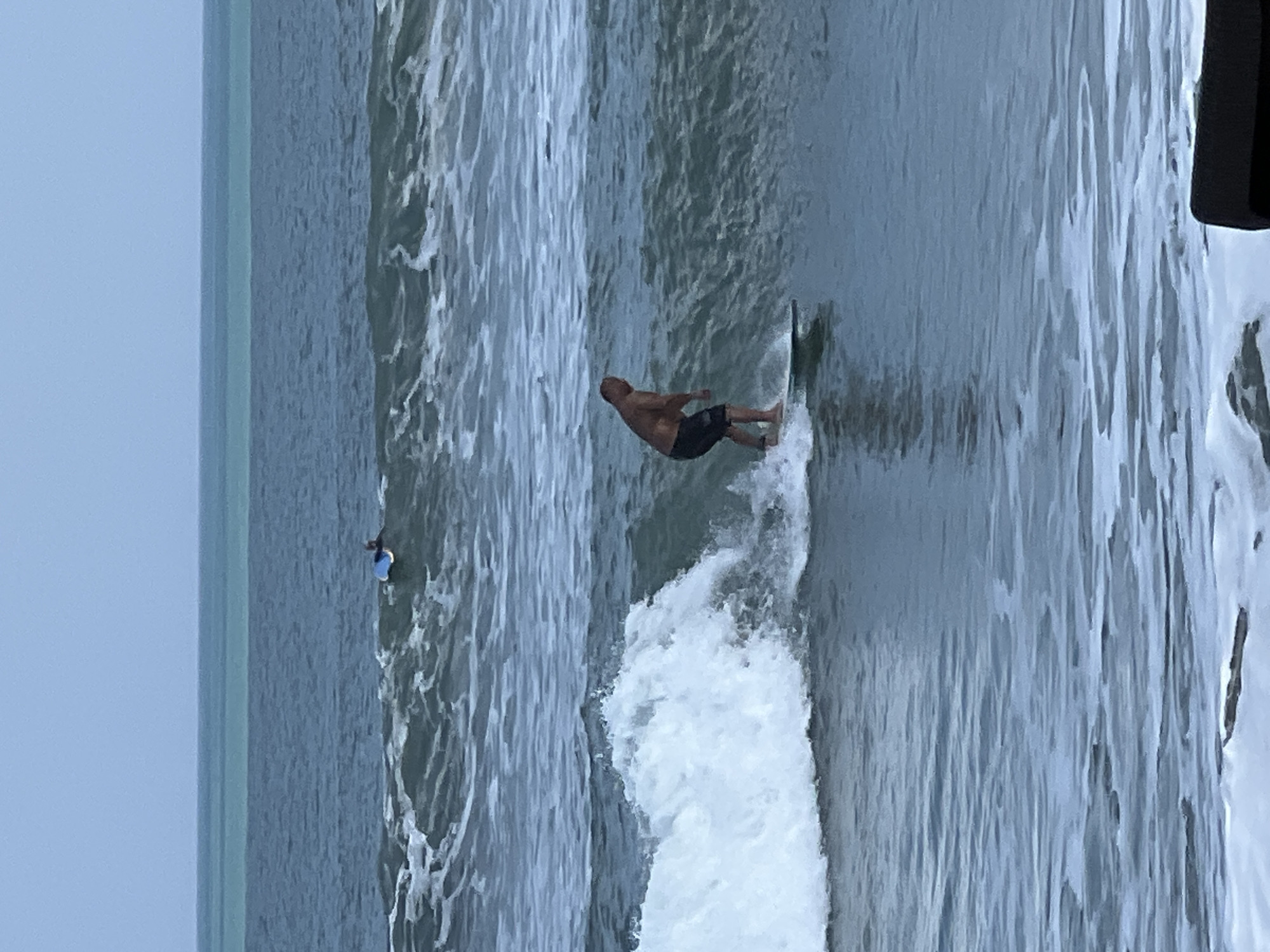 A medium shot captures a man surfing on a wave in the ocean. The man, who appears to be middle-aged and bald, is wearing dark-colored swim trunks and is positioned on a surfboard. He is actively engaged in surfing, with his body leaning into the wave. To the left of the frame, a large wave is breaking, creating white foam and spray. In the background, another surfer is visible, lying on their surfboard in the distance. The ocean water is a deep blue-green color, with choppy waves and white foam scattered throughout. The sky is overcast and a muted blue-gray. The image depicts a scene in Kuta, Indonesia, known for its surfing beaches.