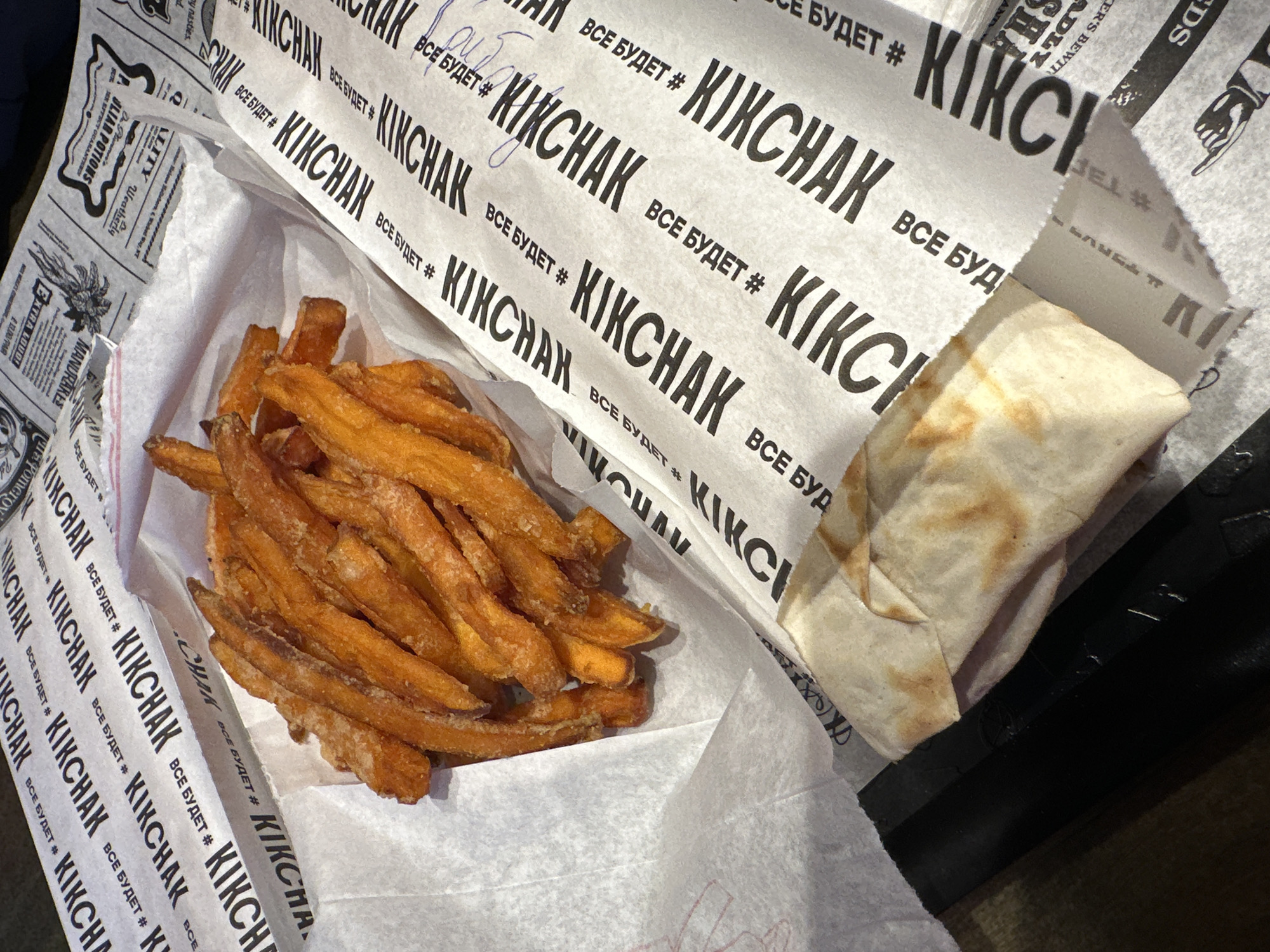 The image displays an elevated view of food items on a branded paper surface, located in Rostov-on-Don, Russia. A white paper container in the upper-left quadrant holds a quantity of irregularly shaped, golden-orange sweet potato fries, visibly coated with crystalline granules. To the lower-right of the fries, a partially unwrapped, beige-colored food item, consistent with a wrap or rolled sandwich, shows light browning and toasted marks on its surface.
The food items are positioned on a white paper sheet featuring a repeating pattern of "KIKCHAK" in large, black, sans-serif capital letters. Below each instance of "KIKCHAK," smaller Cyrillic text reads "ВСЕ БУДЕТ #". Other sections of the paper exhibit faint, newspaper-style printing with English text such as "EXTRA COLD" and "POPULAR POTIONS," alongside various illustrations including a pointing hand and a stylized winged creature. Blue ink markings, resembling a scribble or signature, are visible on the printed paper in the upper-right section. The entire arrangement rests on an indistinct dark surface. No individuals are present in the scene.