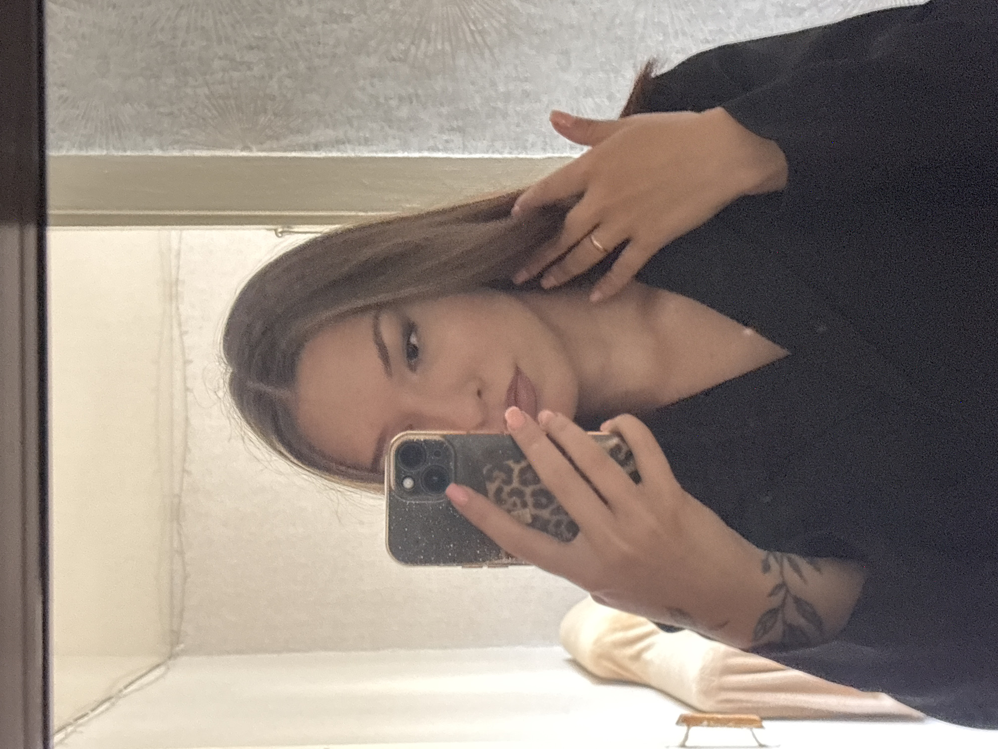 A young woman with long brown hair is taking a selfie in a mirror. She is wearing a black V-neck top. Her left hand is holding a smartphone with a leopard print case. Her right hand is touching her hair. There are tattoos on her left arm. The background appears to be an indoor setting, possibly a room in a house or apartment. The location is not specified in the provided information.