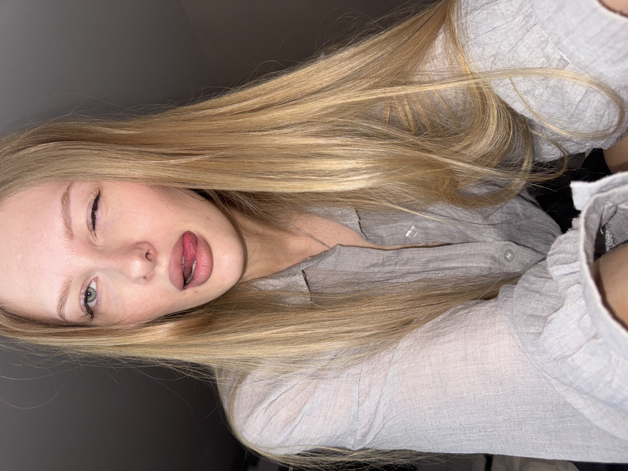A young Caucasian woman with long blonde hair and light-colored eyes is shown in a close-up shot, winking and sticking her tongue out. She is wearing a light gray button-up shirt. The background is dark and appears to be indoors, possibly a room in a residential building, with no discernible city features of Perm, Russia visible.