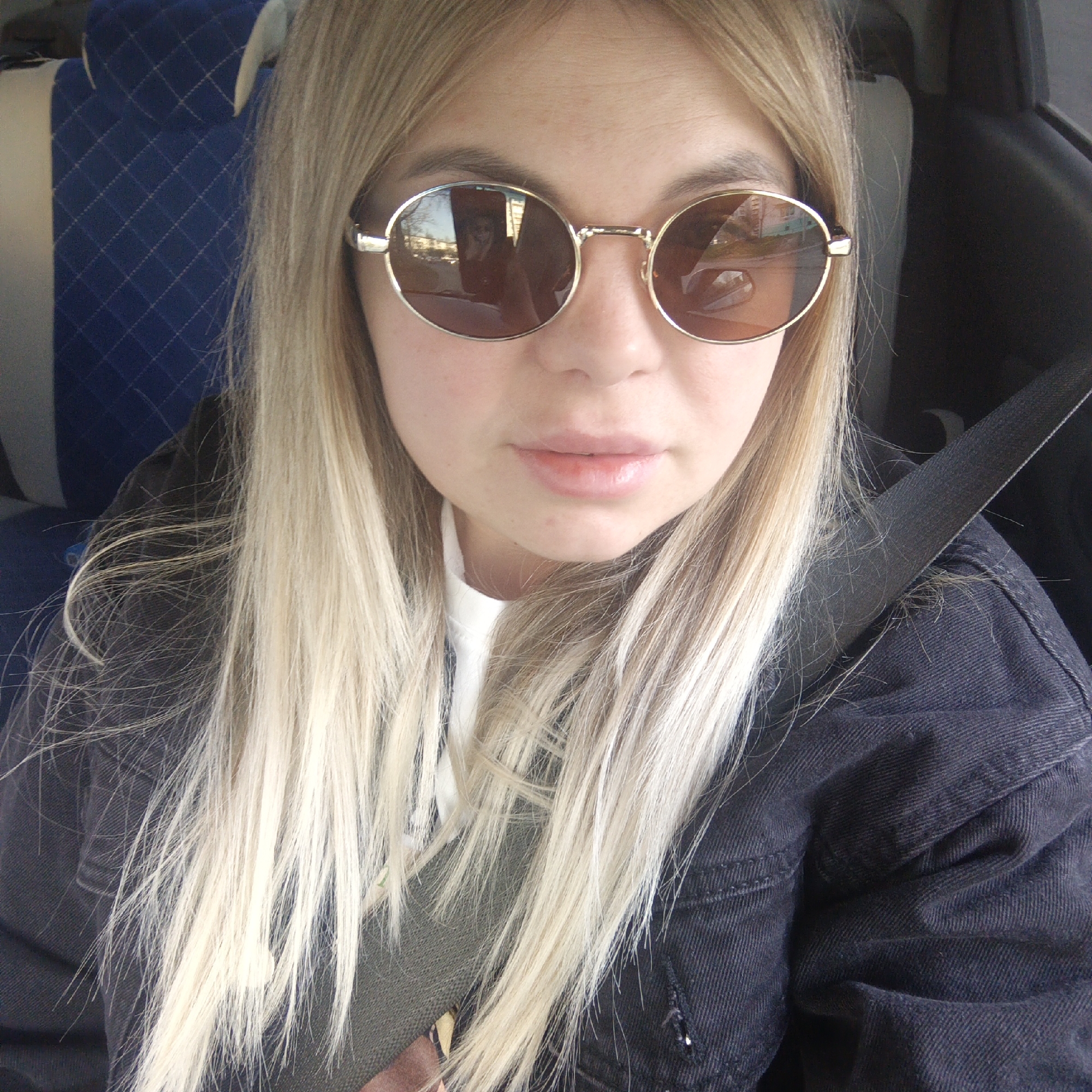 A young woman with long blonde hair is in a car, wearing round gold-framed sunglasses and a black denim jacket. She has on her seatbelt. The car has blue upholstered seats with a quilted pattern. The reflection in her sunglasses shows buildings, possibly indicating an urban setting. Location: Unknown, Unknown.