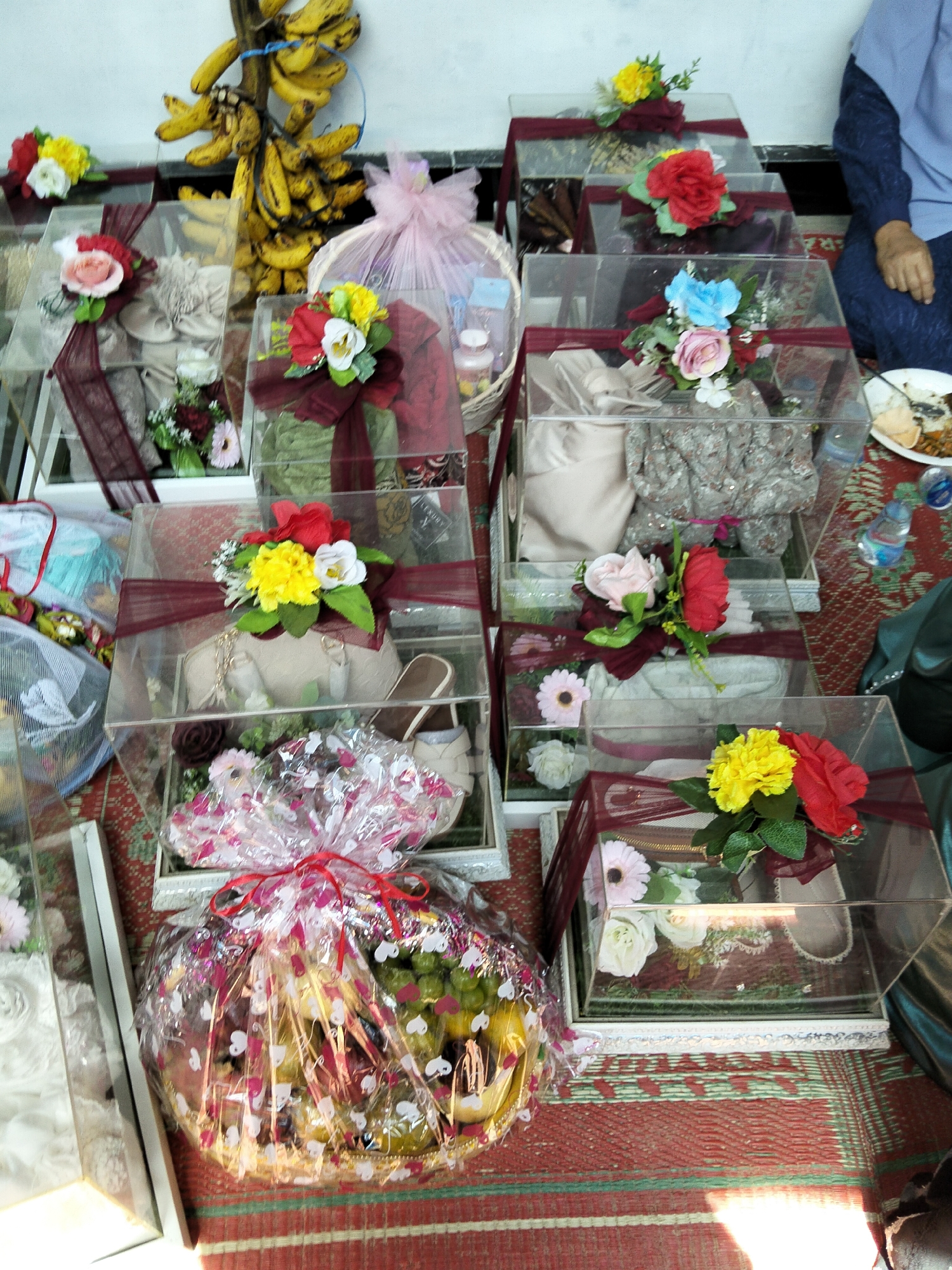 The image displays a collection of gift boxes, likely prepared for a wedding or special occasion, arranged on a patterned rug. The boxes are made of clear acrylic and decorated with maroon ribbon and artificial flowers in various colors, including red, pink, yellow, and white. Some boxes contain fabric items, possibly clothing or veils, while others hold what appears to be a handbag and shoes. In the background, a bunch of bananas hangs from a rack. A person wearing a blue garment is partially visible on the right side of the frame, and a plate with food and a bottle of water are also present. The overall setting suggests an indoor event or preparation area. The specific location is identified as Pulonga, Indonesia.