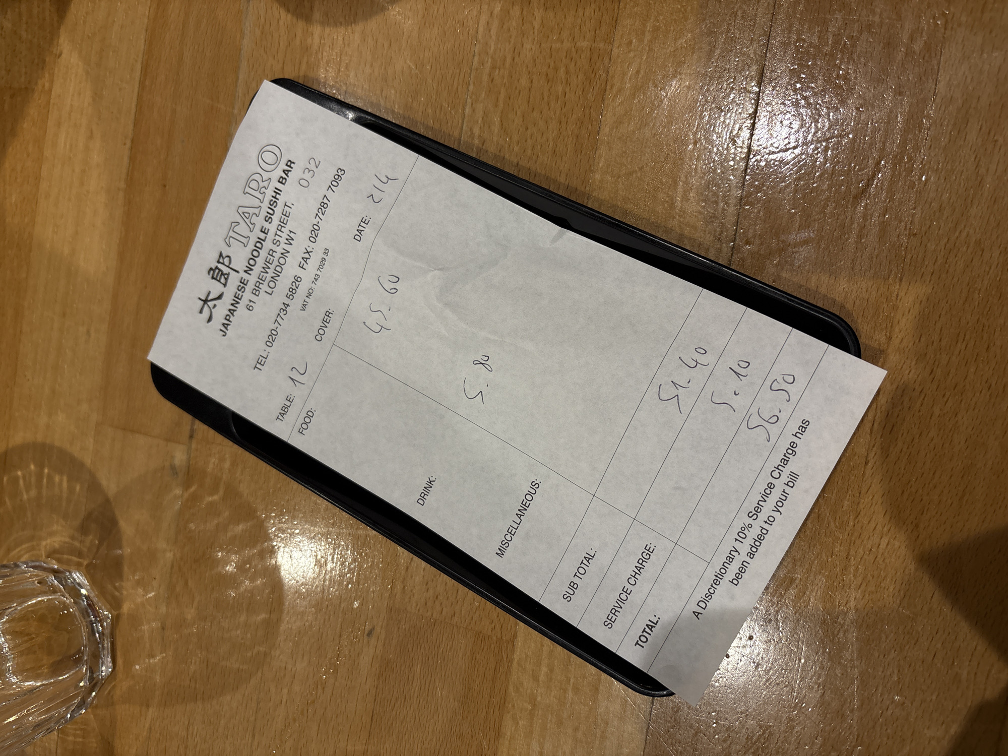 A close-up shot shows a Japanese noodle sushi bar receipt on a wooden table. The receipt is from 大郎 Taro, a Japanese noodle and sushi bar located at 61 Brewer Street, London W1. The table number is 12, and the date is 2/14. The charges are £45.60 for food and £5.80 for drinks. The subtotal is £51.40, the service charge is £5.10, and the total is £56.50. A note at the bottom of the receipt states that a discretionary 10% service charge has been added to the bill. A drinking glass is visible on the bottom left of the shot.
