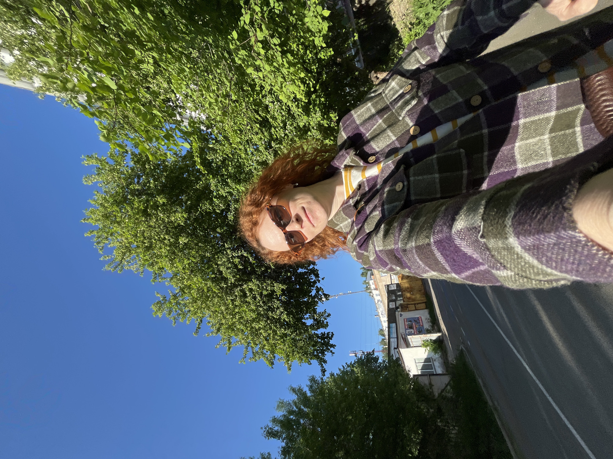 A person with red hair and sunglasses is taking a selfie on a sunny day in Chaikovsky, Russia. The subject is wearing a plaid shirt with a striped shirt underneath. The background consists of a blue sky, green trees, a street with a white line, and buildings. The location appears to be near a road in a residential or commercial area of the city.