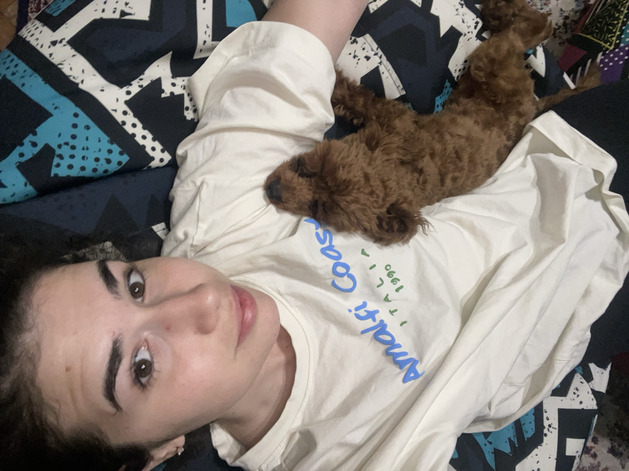 A young woman lies on her back, her face tilted slightly toward the camera. She has fair skin, dark eyebrows, and brown eyes. She wears a light-colored t-shirt with the words "Amalfi Coast Italy 1990" printed on it in blue and green. A small, brown poodle-type dog lies on her chest. The background consists of a patterned blanket or duvet cover in shades of blue, black, and white. The location cannot be determined from this image.