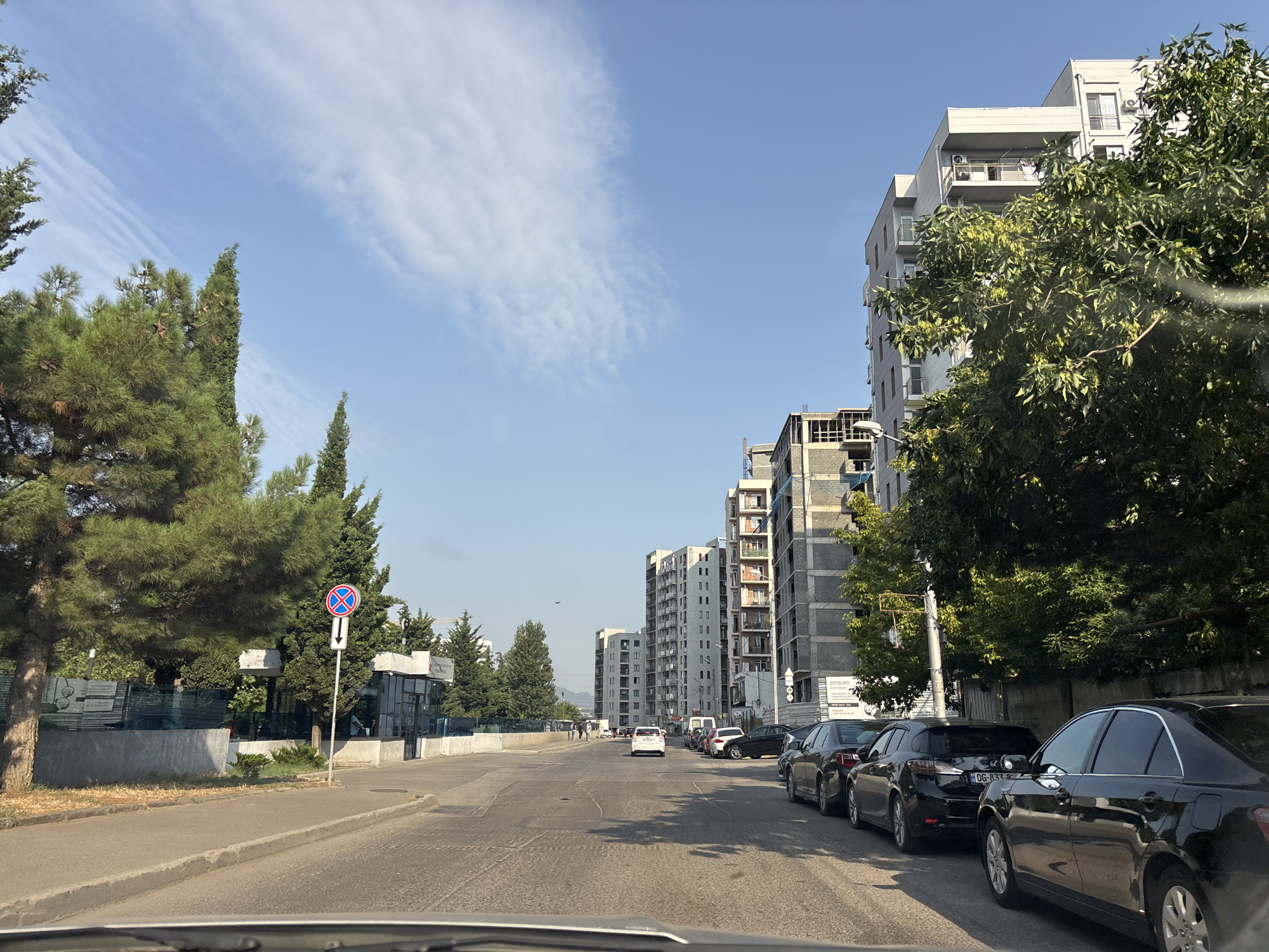 The image depicts a street scene in Tbilisi, Georgia, on a clear, sunny day. The perspective is from within a vehicle, looking forward down a paved road. On the right side of the road are several multi-story residential buildings, some of which appear to be under construction, with concrete structures and exposed rebar. Lined up along the curb are parked cars, mostly black sedans and SUVs. Lush green trees, including pine trees, line the left side of the road, providing shade. A blue traffic sign with a red circle and a diagonal red line, indicating no stopping, is visible near the trees. Beyond the trees, a low, modern-looking building with glass panels is situated. In the distance, more apartment buildings are visible, and the sky is a clear blue with wispy white clouds. A few pedestrians are seen walking on the sidewalk in the middle distance. The overall impression is one of urban development with a mix of residential buildings, ongoing construction, and natural elements.