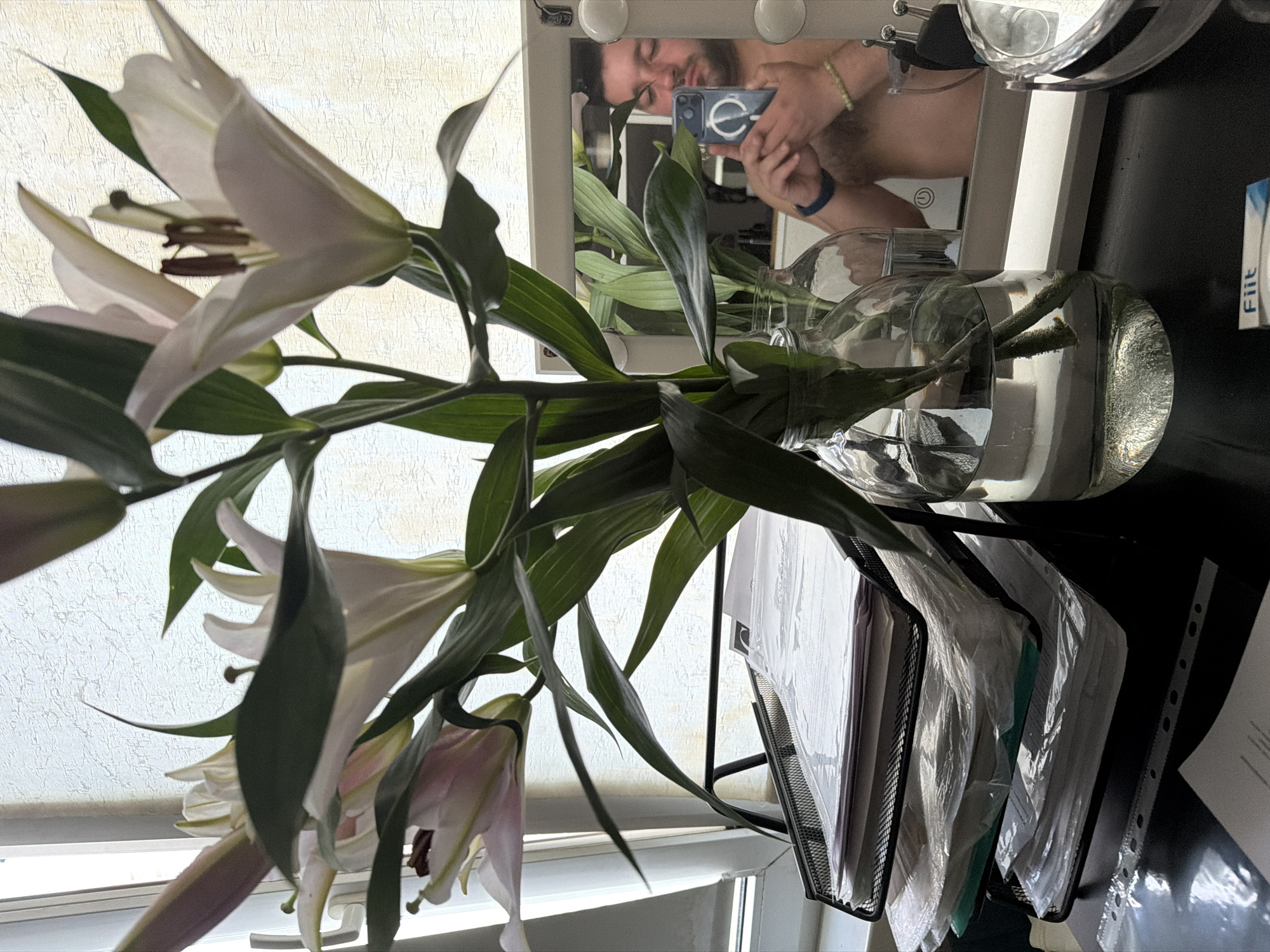 A man is reflected in a bathroom mirror, taking a picture with his phone. He has a beard, and he is bare-chested. In front of the mirror, there is a glass vase filled with water and lilies. To the right of the vase, there is a black file organizer with papers. The room appears to be well-lit, with a window behind the flowers. The location is not identified in the city of Moscow, Russia.
