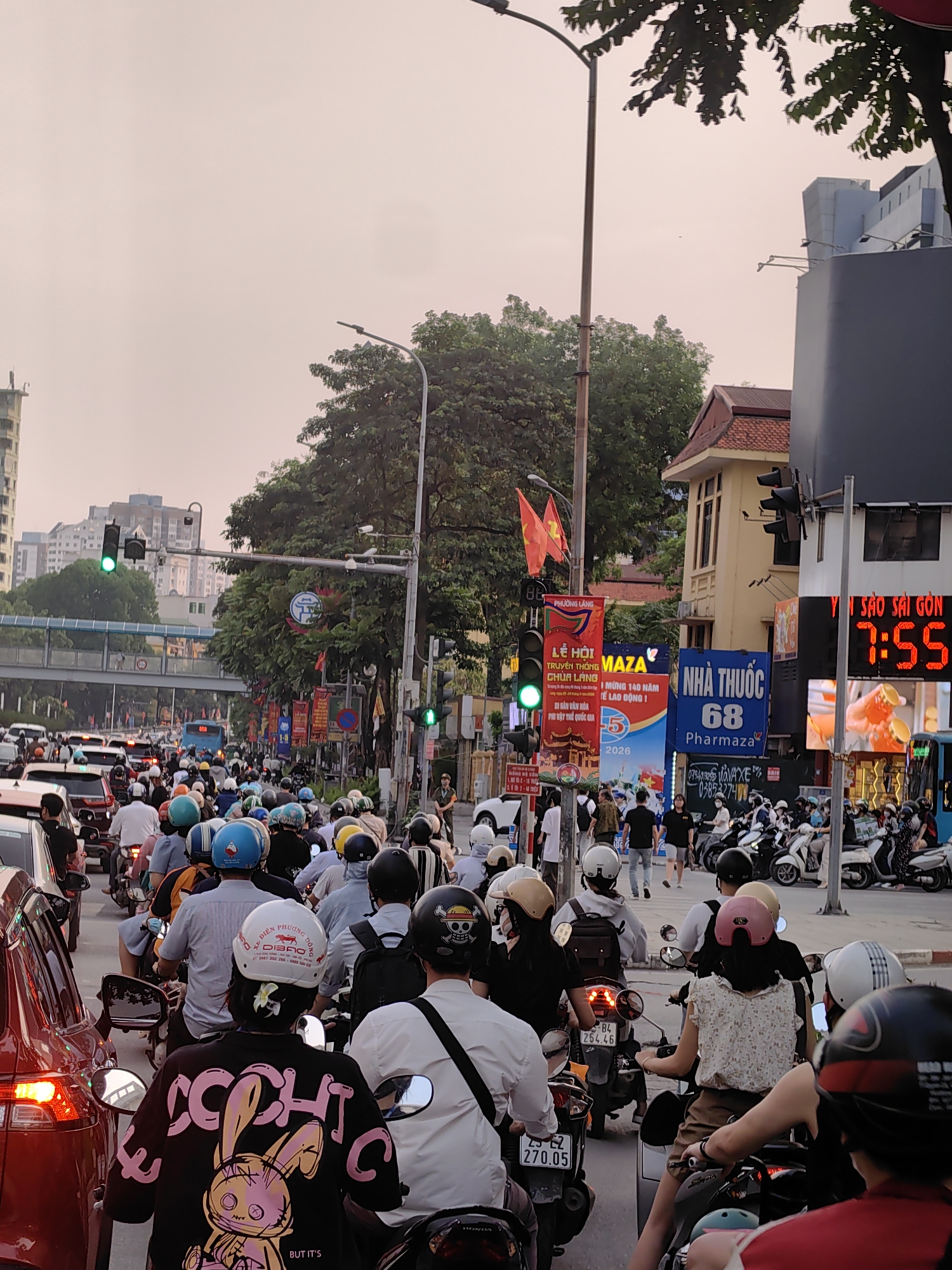 The image captures a bustling street scene in Hà Nội, Vietnam, characterized by heavy traffic. Numerous people on motorbikes dominate the foreground and mid-ground, many wearing helmets of various colors and designs. A red car is visible in the left foreground, caught in the same congestion.
The setting is an urban environment, with multi-story buildings, dense green trees, and streetlights. An overpass or pedestrian bridge can be seen in the background. The sky is overcast or hazy, providing a soft, diffused light that suggests either early morning or late afternoon. A prominent digital clock on the right side of the image displays "7:55," accompanied by the text "Y SÁO SAI GÒN" (Bird's Nest Saigon).
The street is adorned with several signs and banners. A large blue sign advertises "NHÀ THUỐC 68 Pharmaza" (Pharmacy 68 Pharmaza). Other banners promote "LỄ HỘI TRUYỀN THỐNG CHÙA LÁNG" (Traditional Festival Chua Lang) and "MỪNG 140 NĂM TẾ LAO ĐỘNG 1" (Celebrating 140 Years of Labor Day 1), with "PHƯƠNG LÁNG" and "MAZA" also visible. Two Vietnamese national flags (red with a yellow star) are mounted on a pole.
Specific details include text on helmets, such as "DIỄN PHƯƠNG D" and "DIBRO" on a white helmet, and a black jacket with a pink rabbit graphic and "BUT IT'S" printed below. Vehicle license plates, like "84 254.46" and "29 E2 270.05," are discernible. Green traffic lights are visible, indicating either waiting traffic or just-released lanes. The overall impression is one of a vibrant, busy city, characteristic of Vietnamese urban life.
