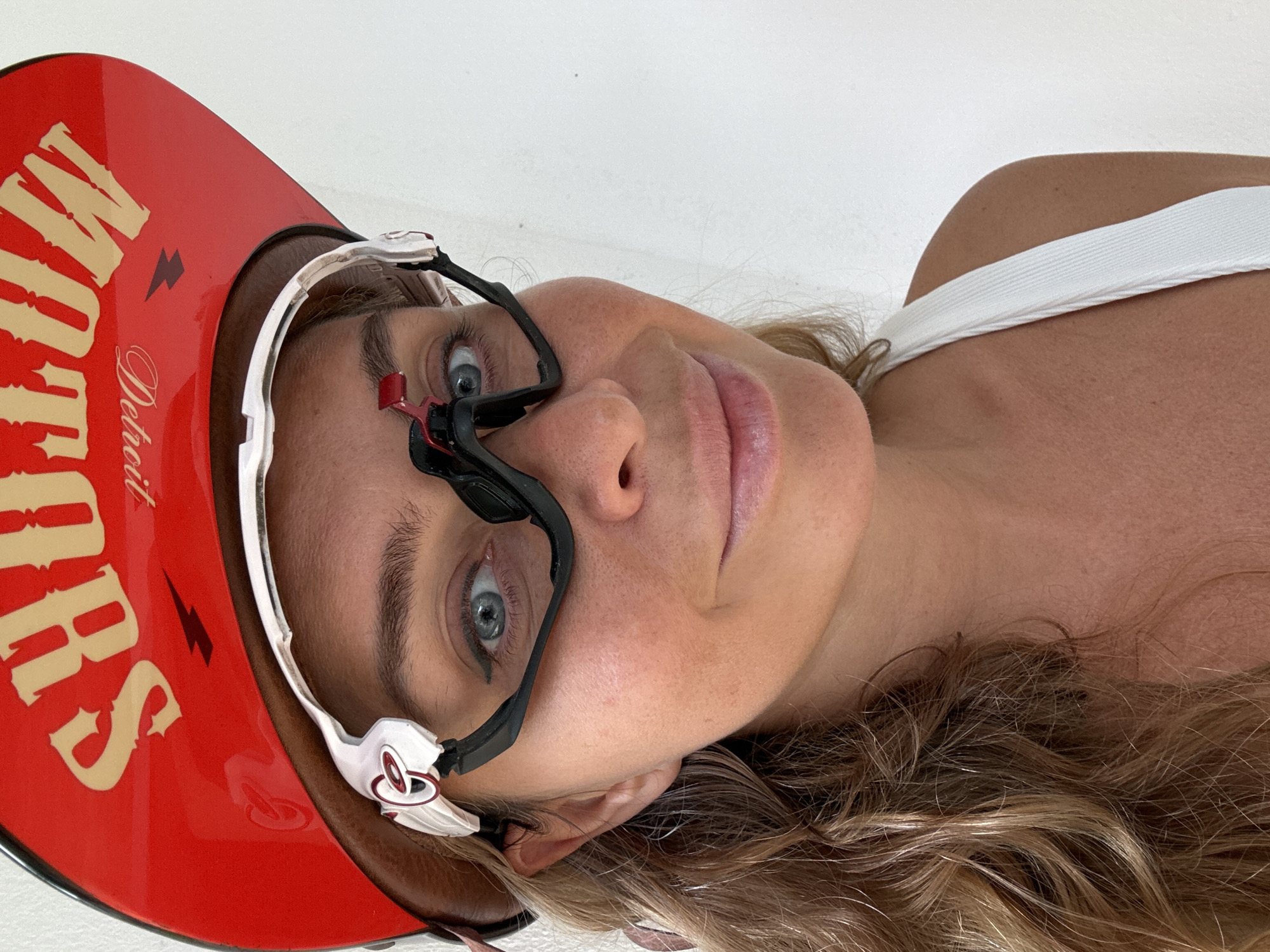 A close-up, slightly high-angle medium shot shows a woman with blonde hair wearing a red hat with gold lettering that says "MOTORS DETROIT" and black and white goggles. She is looking directly at the camera with a slight smile. She is wearing a white tank top. The background is a plain white wall. The image was taken in the city of Ko Samui District, Thailand.