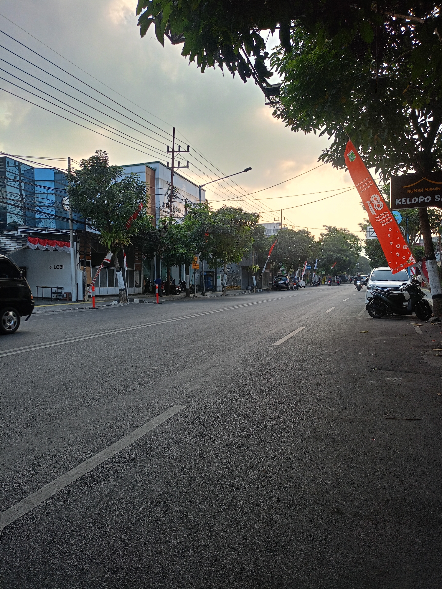 This image depicts a street scene in Batu, Indonesia, during the day, with the sky exhibiting hues of sunset. The road surface is asphalt with a white line marking the lane division. On the left side of the street, there are buildings, including one with a blue glass facade and another with a white facade. A black van is visible on the left, and a sign with the word "LOBI" is present. Trees are planted along the sidewalk.
On the right side of the street, a white car and a black motorcycle are parked. A tall, orange banner with Indonesian text and the number "18" is displayed. Another sign indicates "RUMAH MAKAN KELOPO SA". Several other motorcycles are parked further down the street, and various other vehicles, including cars and motorcycles, are seen on the road. The environment suggests a commercial or urban area with residential elements.