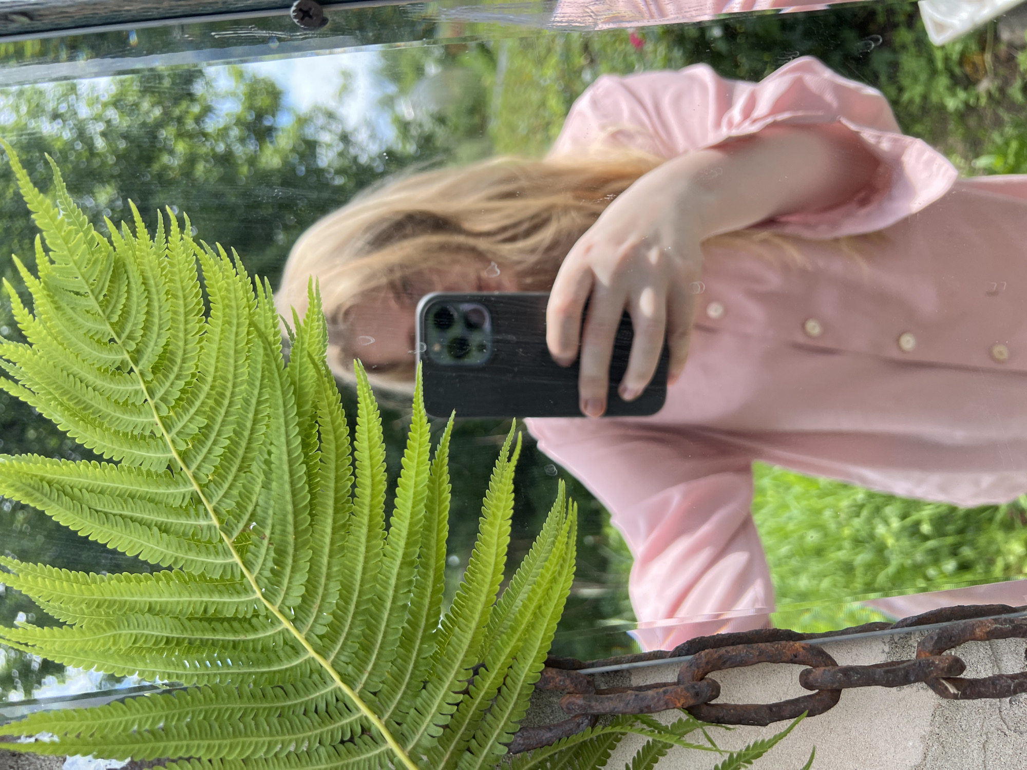In this photograph, a young woman with blonde hair is reflected in a mirror. She is wearing a pink collared shirt with white buttons and is holding a dark-colored smartphone in her right hand, angled to capture her reflection. A large, green fern frond is positioned in the foreground, partially obscuring the mirror. To the left of the fern, a rusty metal chain is attached to a textured concrete surface. The background, visible through the mirror, shows a lush, green garden with trees and foliage, suggesting an outdoor setting. The image does not provide information about the specific location within the city of Chaikovsky, Russia.